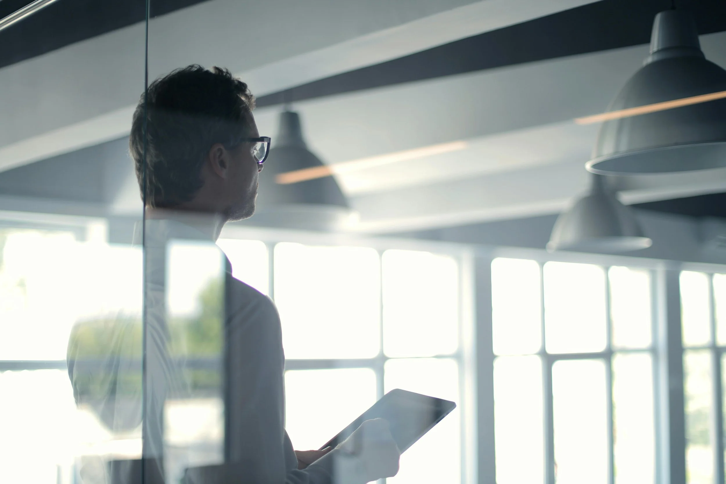 A business professional holding a tablet and looking thoughtfully out the window of a modern commercial office space.