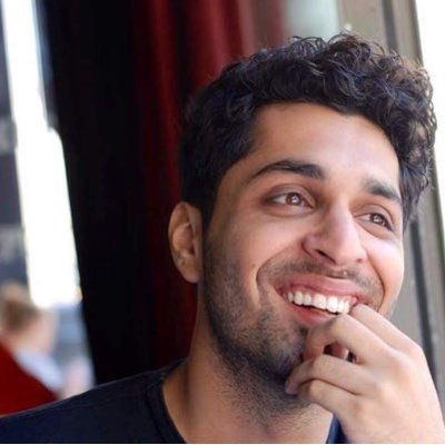 A young man with dark curly hair and a beard, smiling and touching his lips, looking out a window.