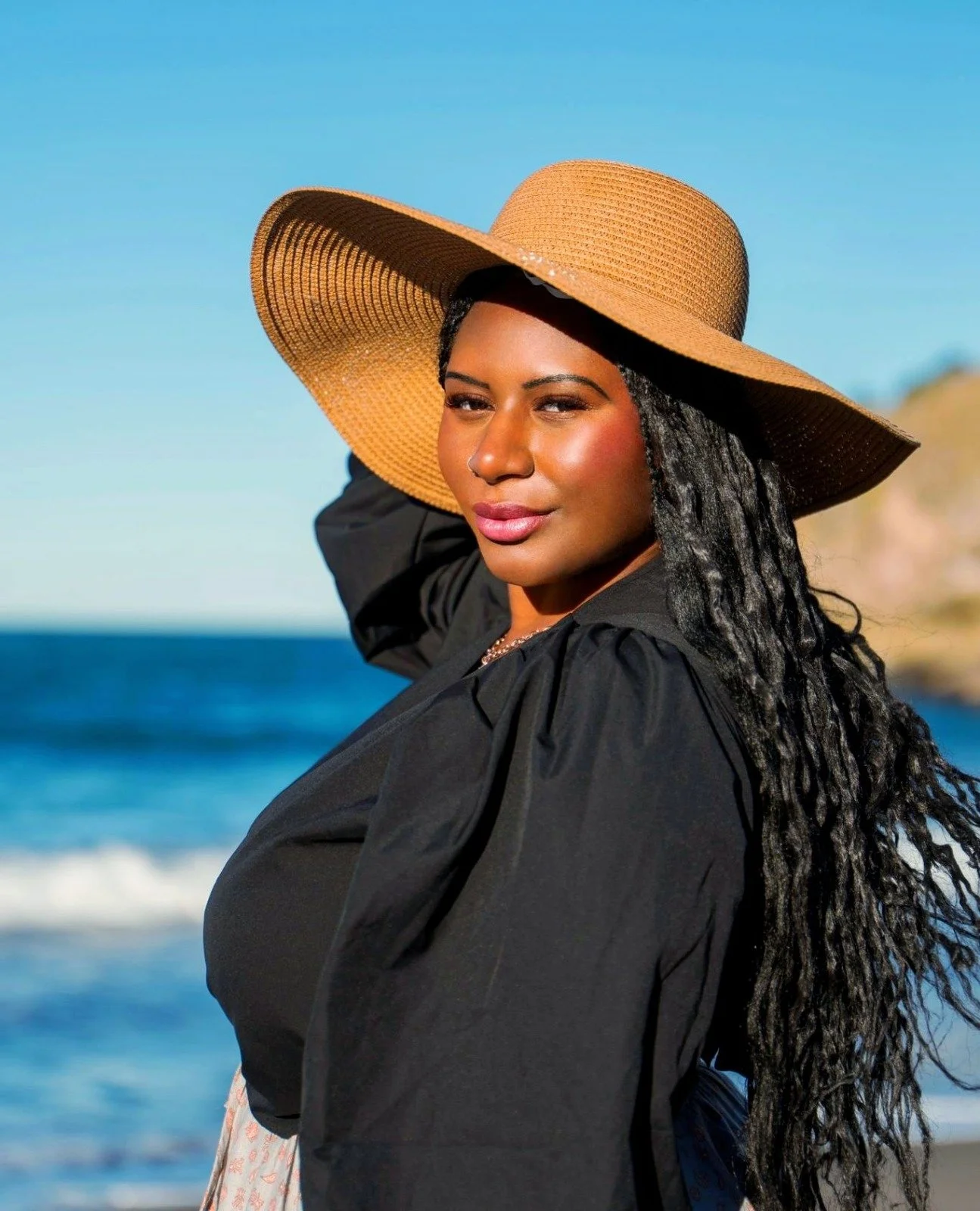 Woman with long braided hair wearing a large straw hat and black clothing standing on the beach with ocean and cliffs in the background.