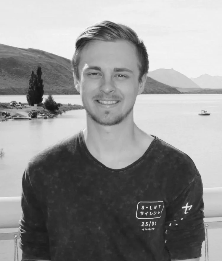 A young man smiling outdoors near a body of water with hills and mountains in the background.