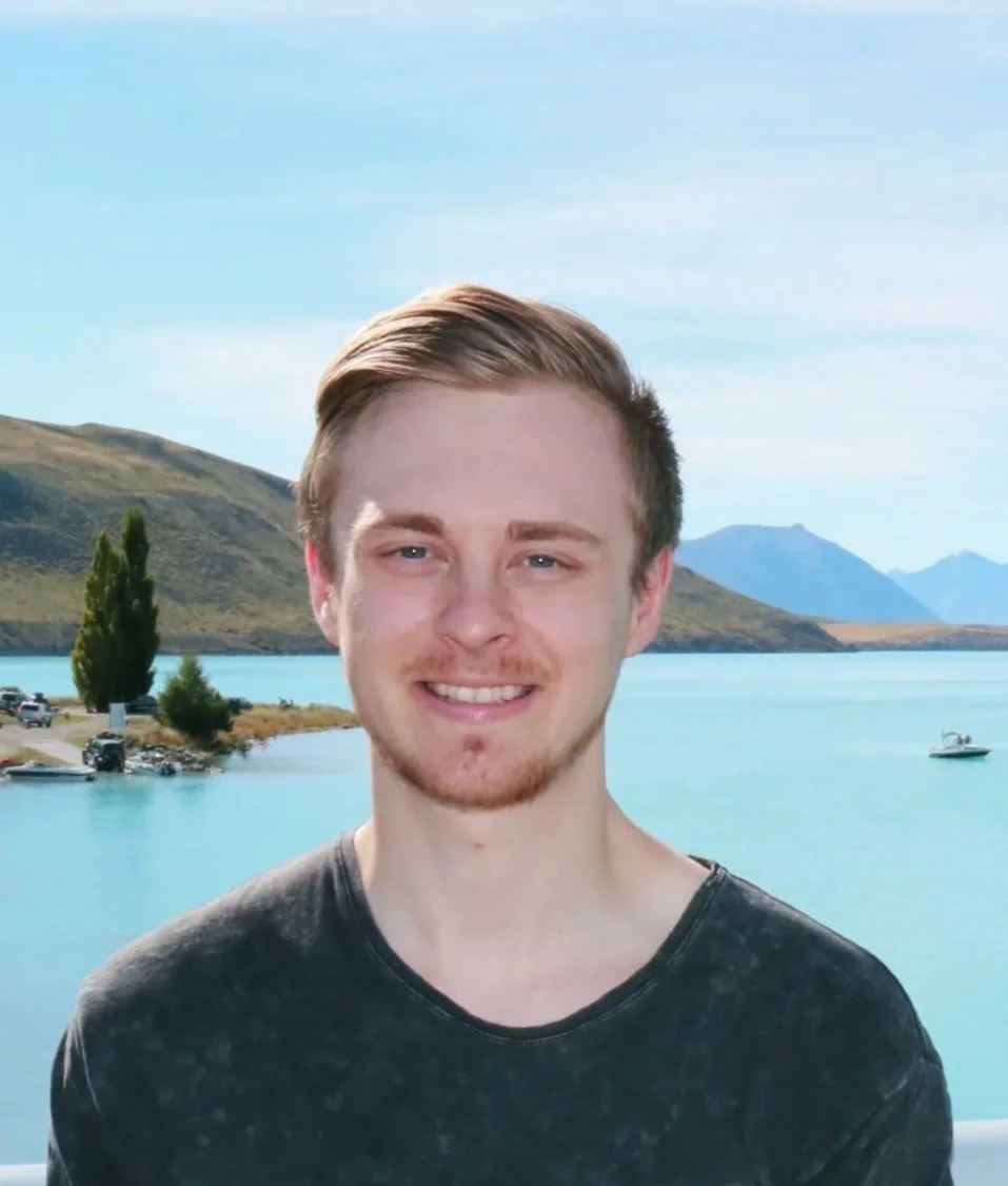 Young man with blond hair and a beard smiling outdoors with a lake, mountains, and a boat in the background.