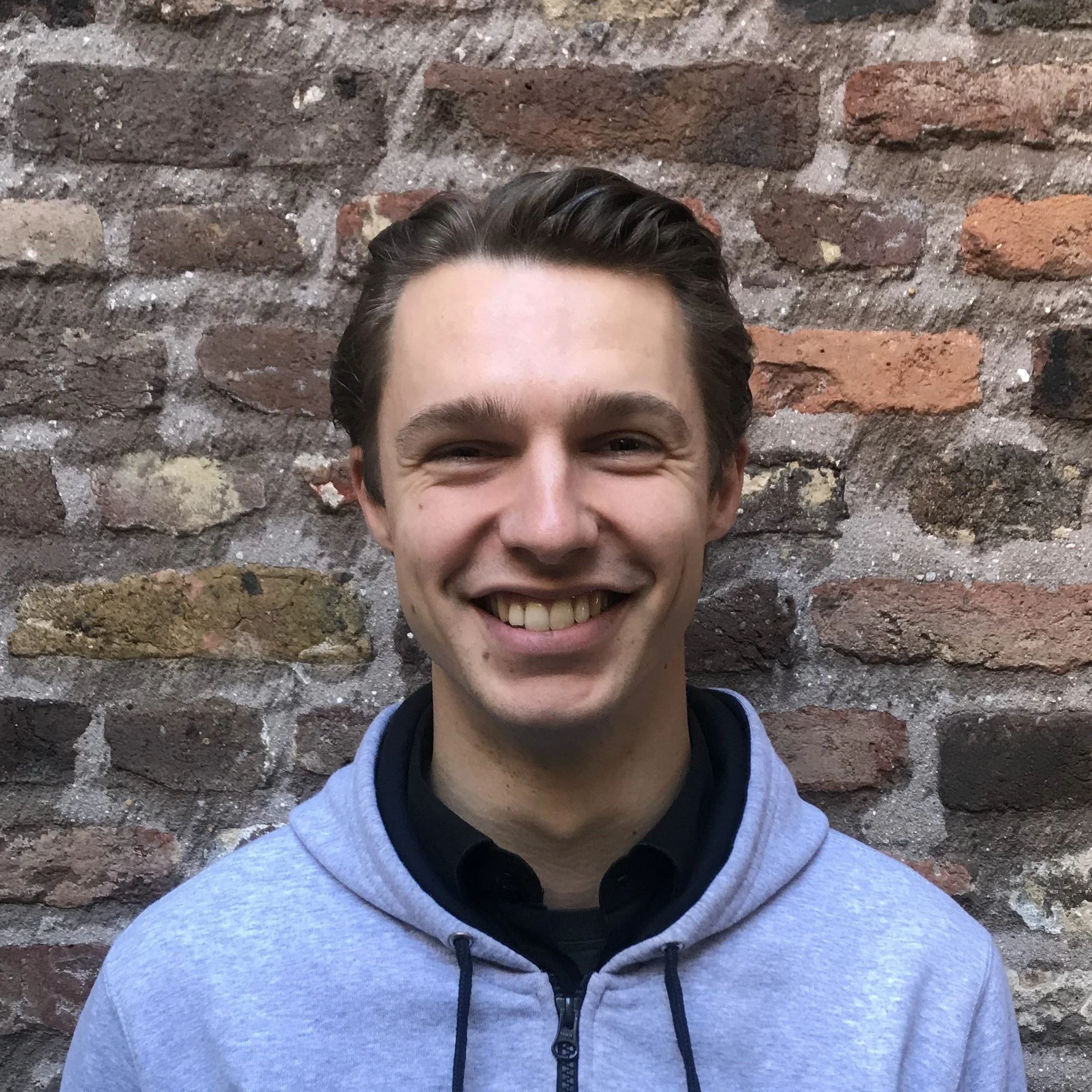 A young man with short dark hair smiling, wearing a gray hoodie over a black shirt, standing in front of a brick wall.