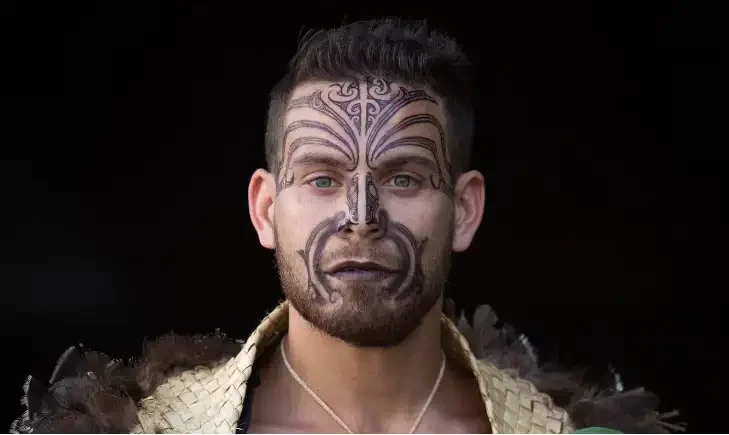 Man with detailed black Maori face paint, wearing traditional clothing, against a dark background.
