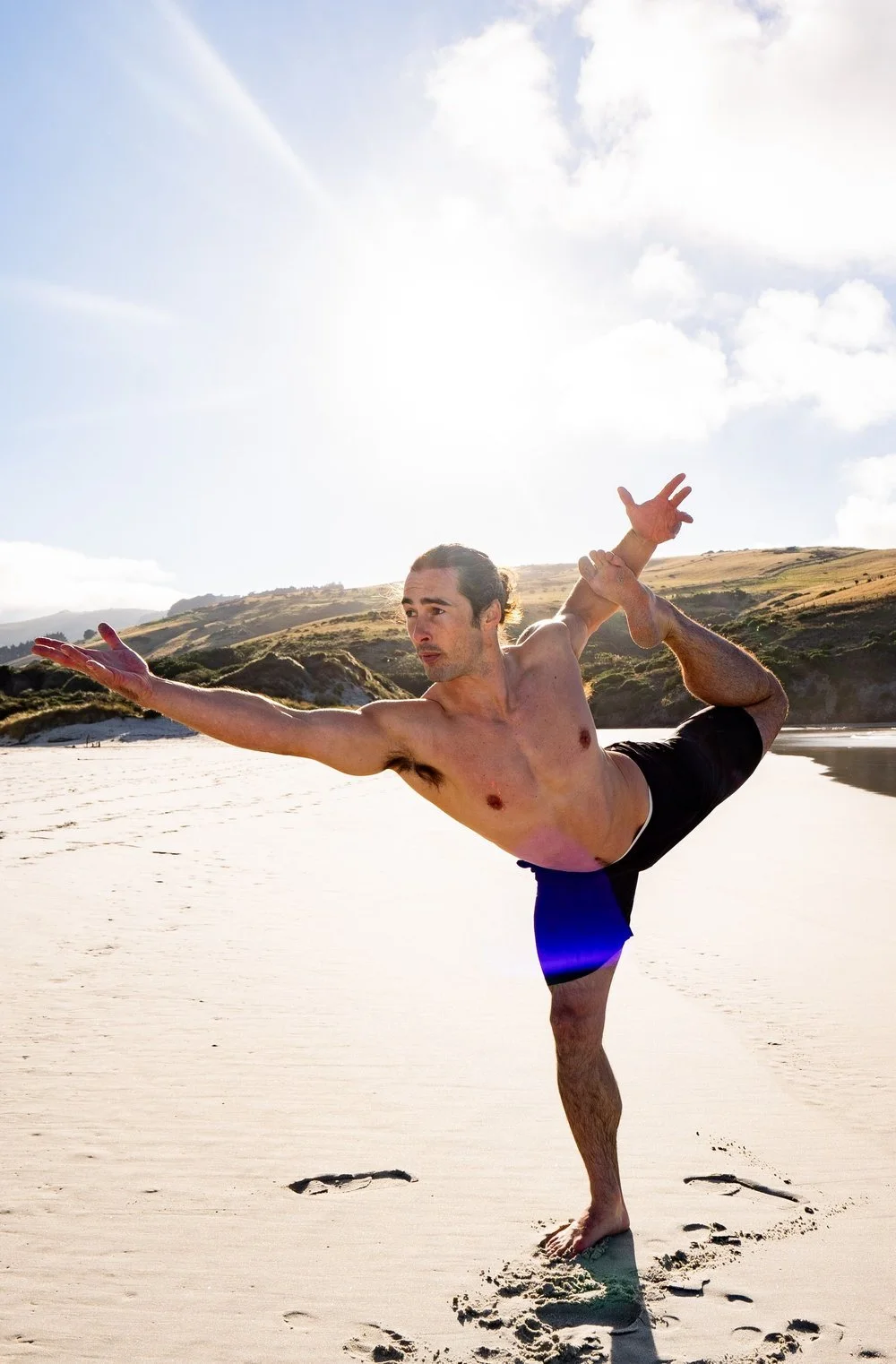A man practicing yoga on a sandy beach with grassy hills in the background, performing a balanced pose with one leg raised behind him and one arm extended forward.