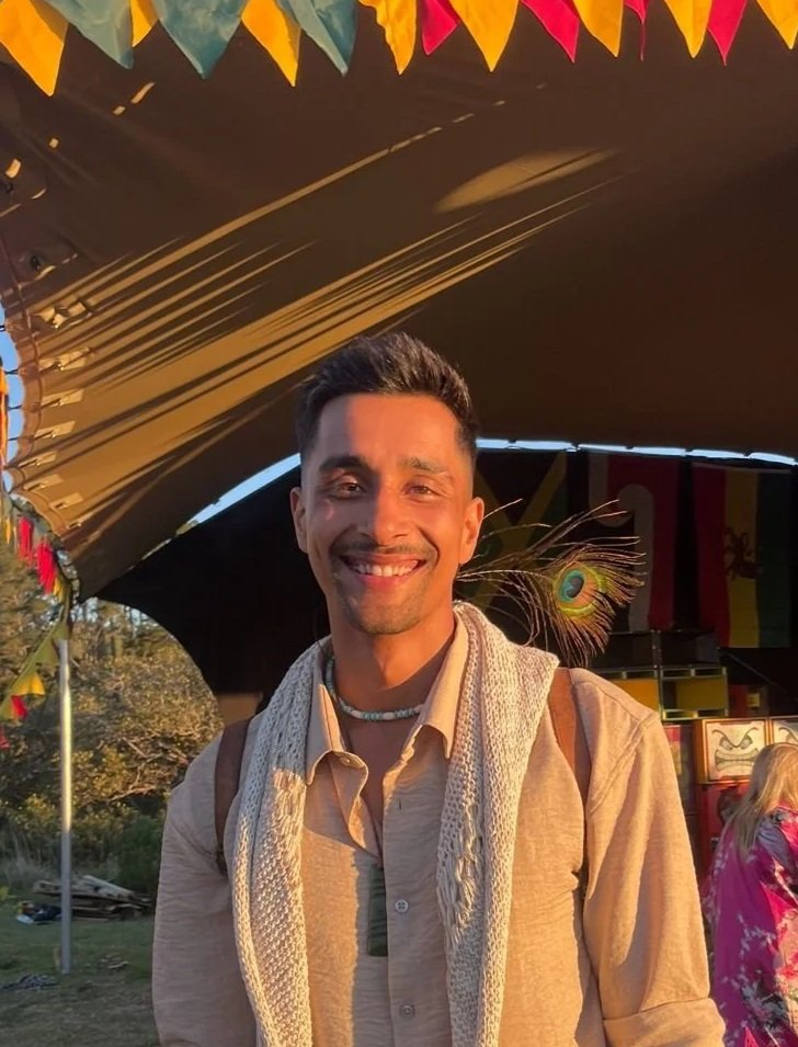 A smiling man with short dark hair, wearing a beige shirt and a white scarf, stands outdoors at a colorful event or festival during sunset, with a large canopy and decorative buntings overhead.