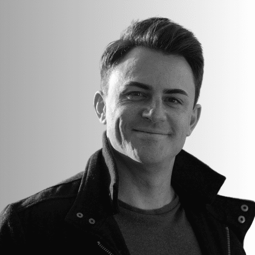 Black and white photo of a smiling young man with short hair, wearing a jacket over a shirt, looking at the camera.