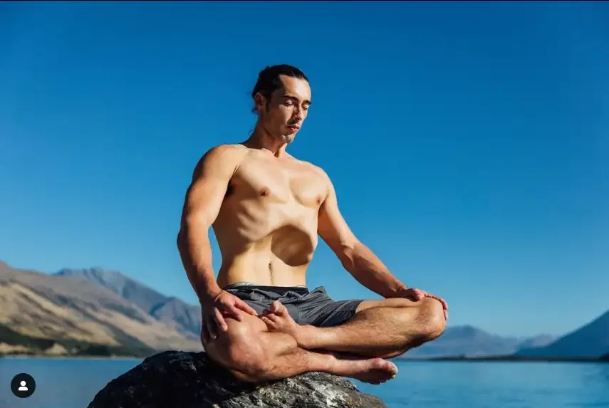 A shirtless man with long hair tied back in a ponytail practicing yoga outdoors, seated in a cross-legged meditation pose on a rock by a lake with mountains in the background under a clear blue sky.