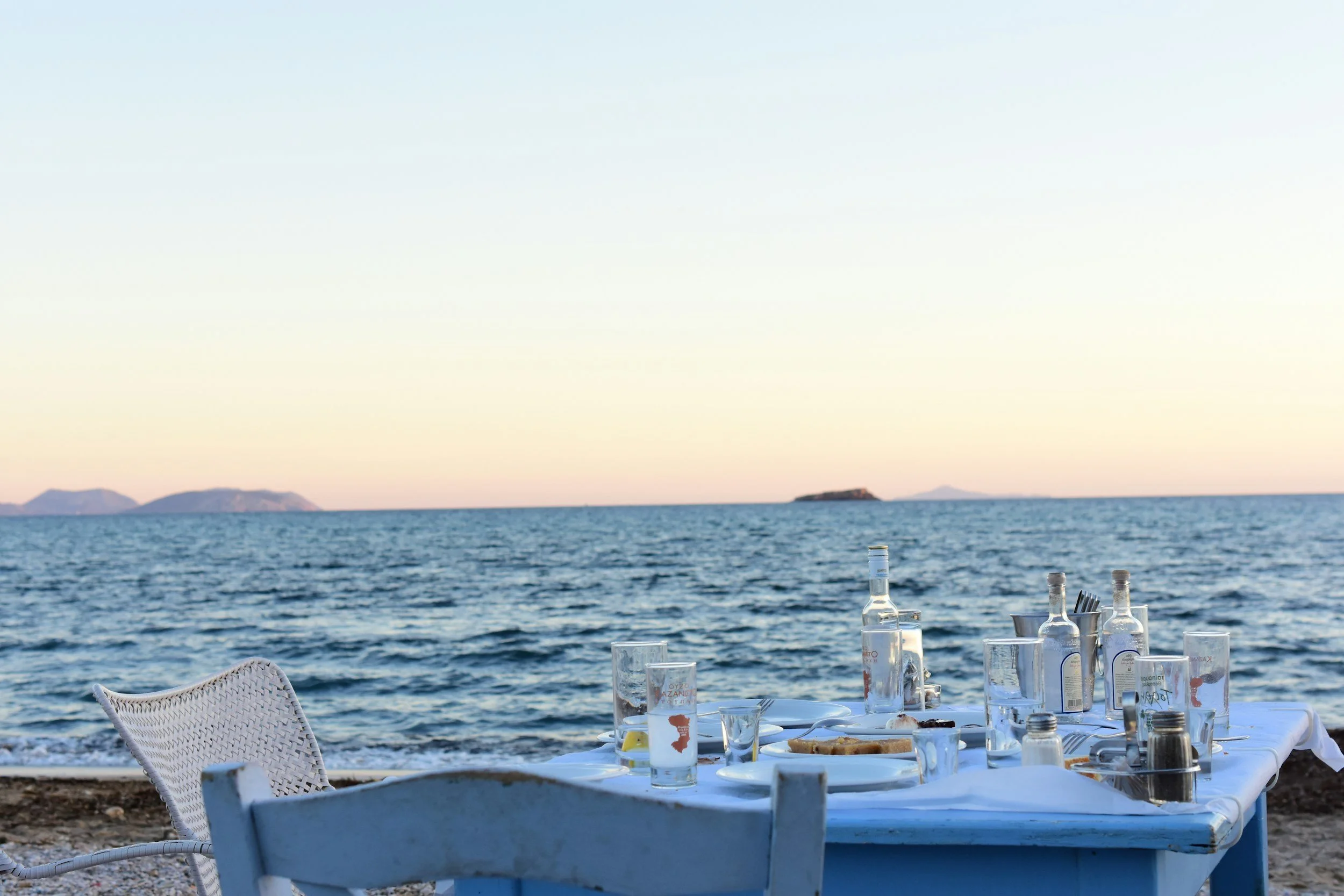 Outdoor dining table set on a beach with ocean and islands in the background during sunset.