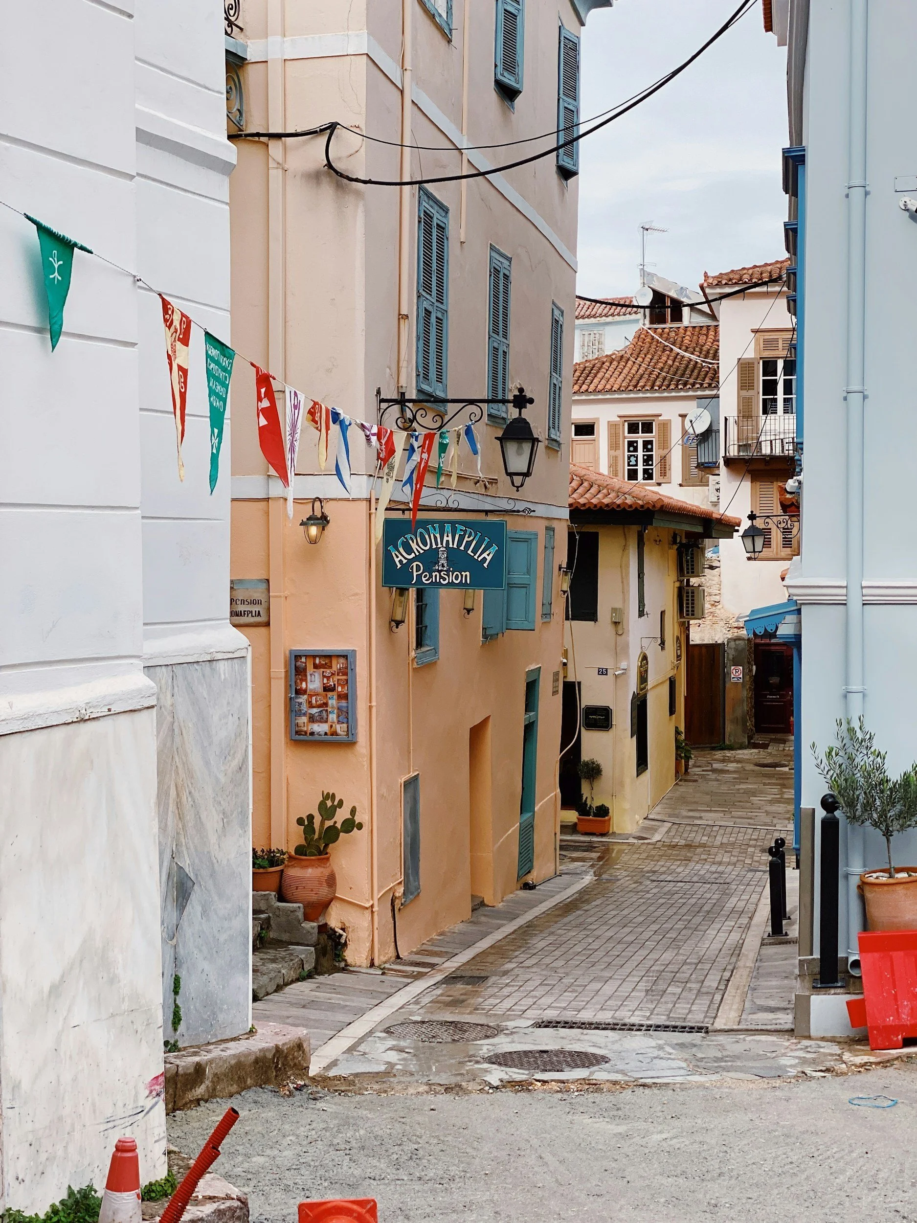 Narrow street in a European town with pastel-colored buildings, blue shutters, hanging flags, potted plants, and a sign for Acropalia Pension.