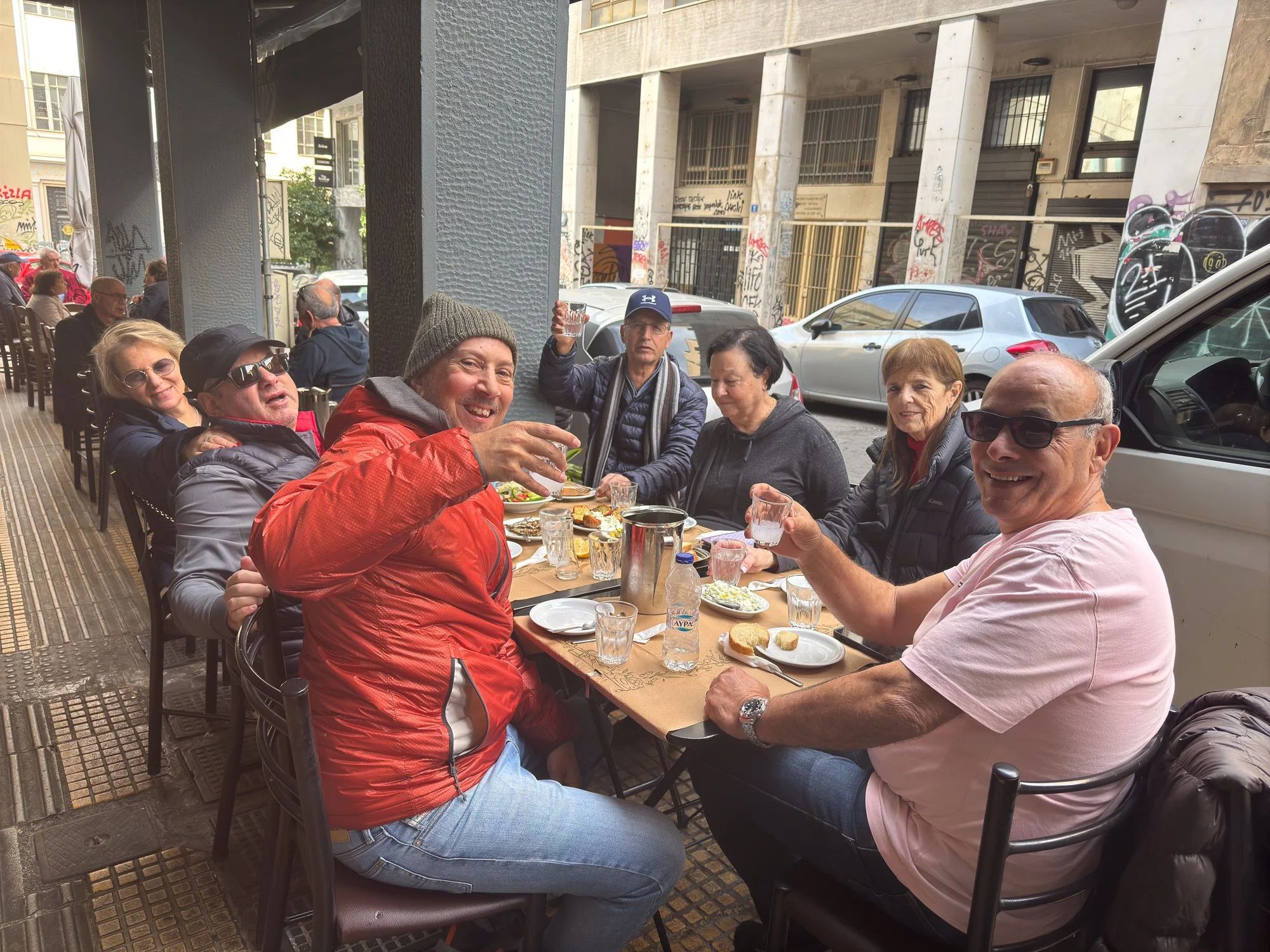 A group of seven people sitting at a long outdoor table at a cafe, enjoying food and drinks, with some raising glasses in a toast, on a city sidewalk with parked cars and graffiti-covered walls in the background.