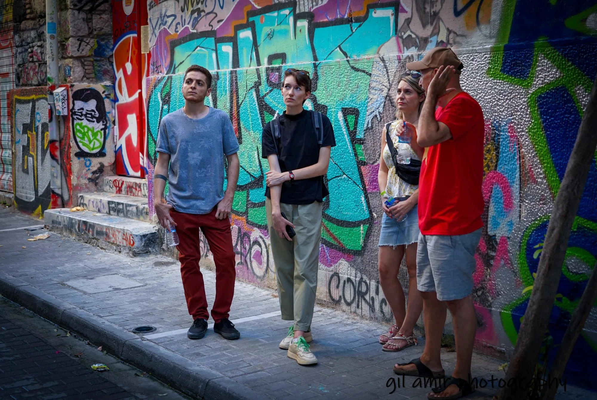 Four people stand in front of a colorful graffiti wall on a city sidewalk, wearing casual summer clothes, holding water bottles and snacks, with one person holding a phone.