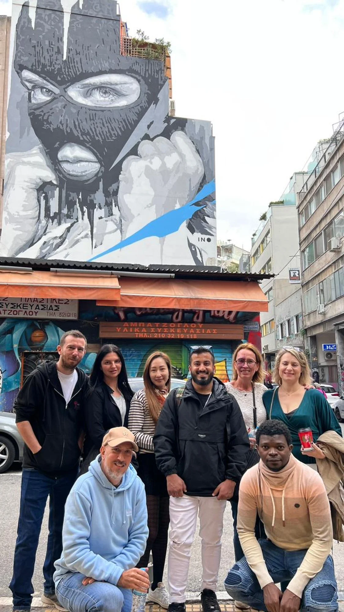 A group of eight people posing in front of a colorful graffiti-covered building with a large mural of a person wearing a ski mask, showing intense eyes and fists, on an urban street.