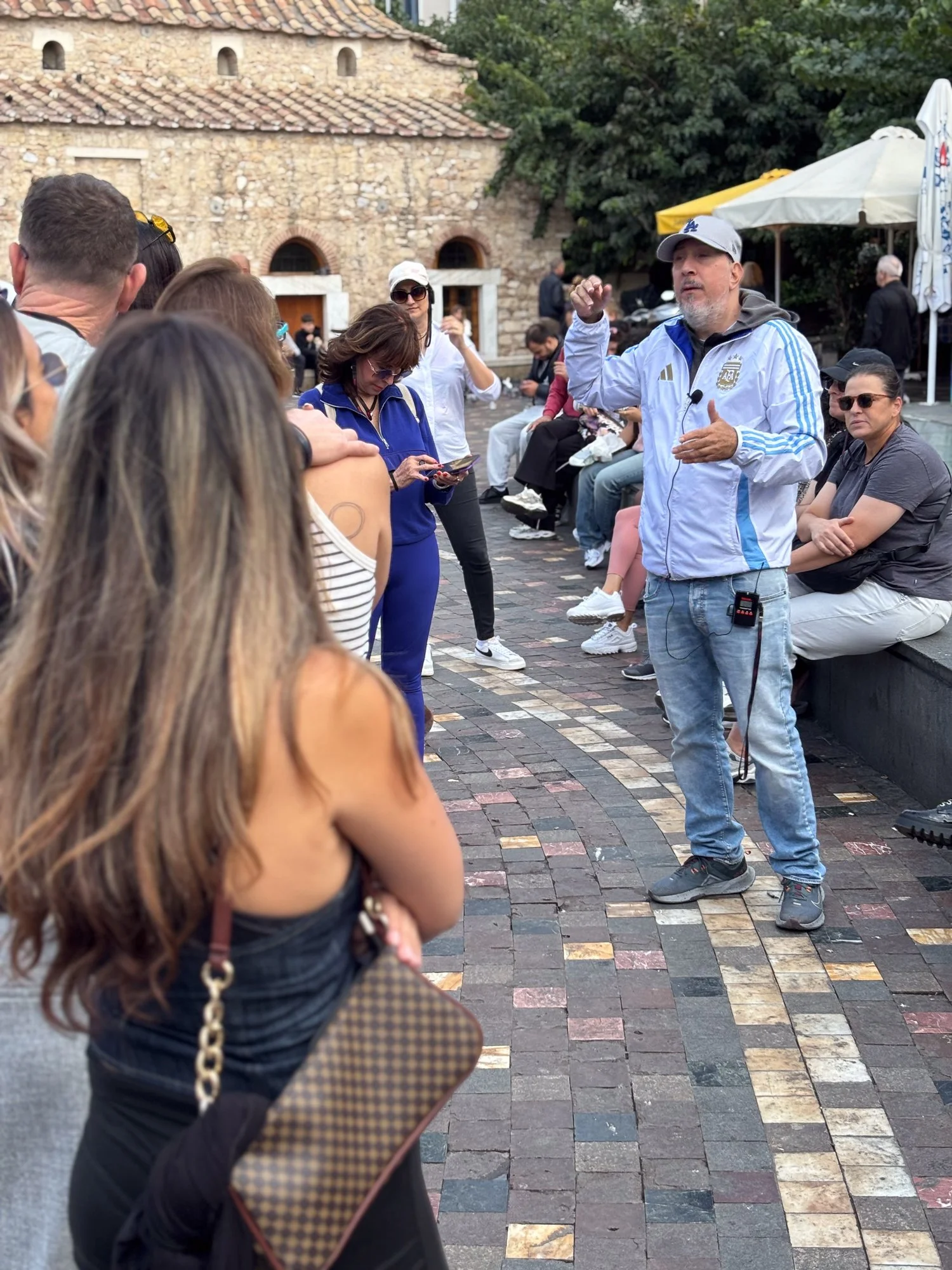 A man giving a speech to a crowd in an outdoor setting with stone building walls in the background, some people sitting on benches, and others standing and listening attentively.