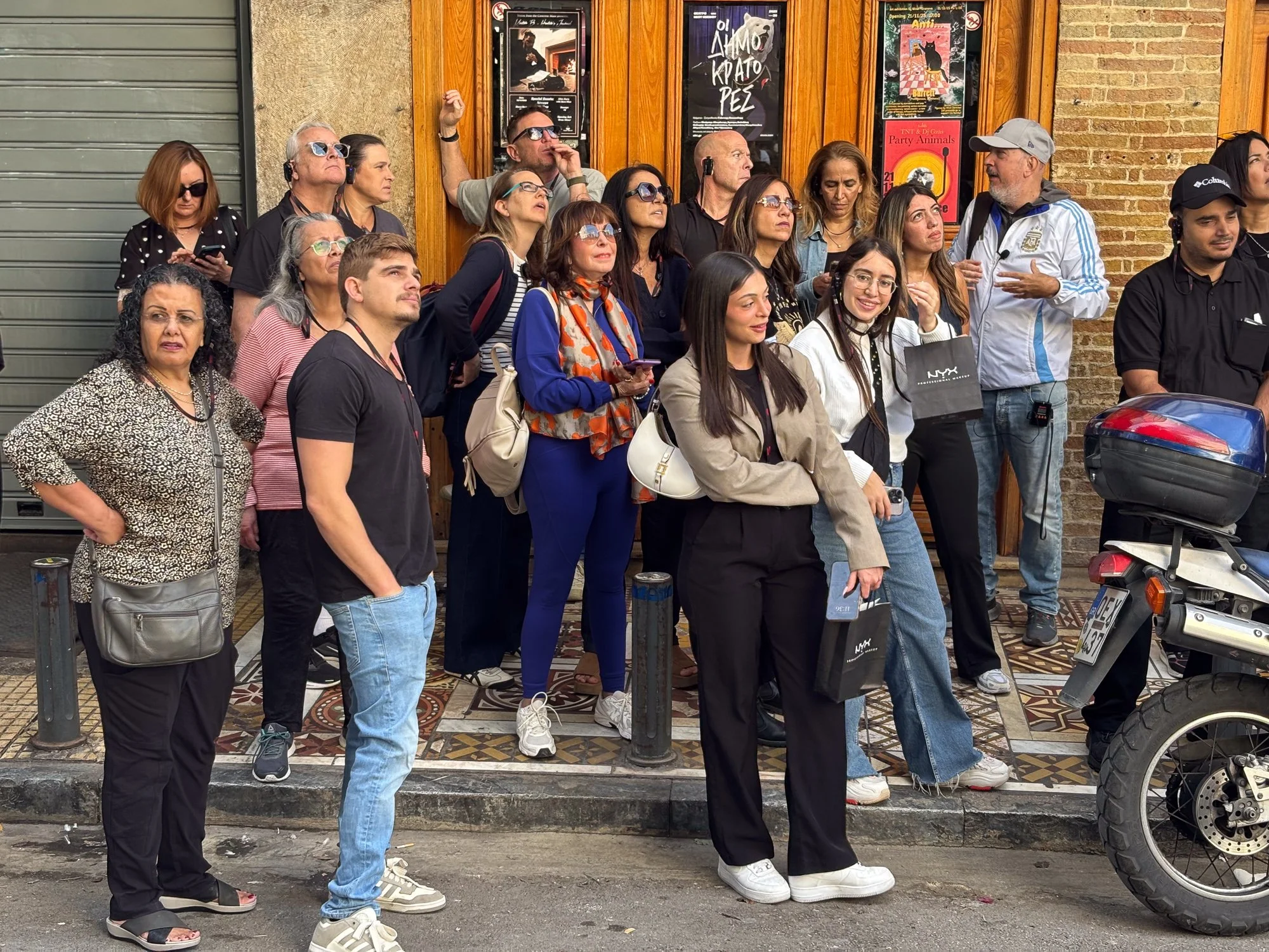 A group of diverse people standing on a city sidewalk outside a building, some wearing sunglasses, with posters on the wall behind them, and part of a motorcycle visible on the right.