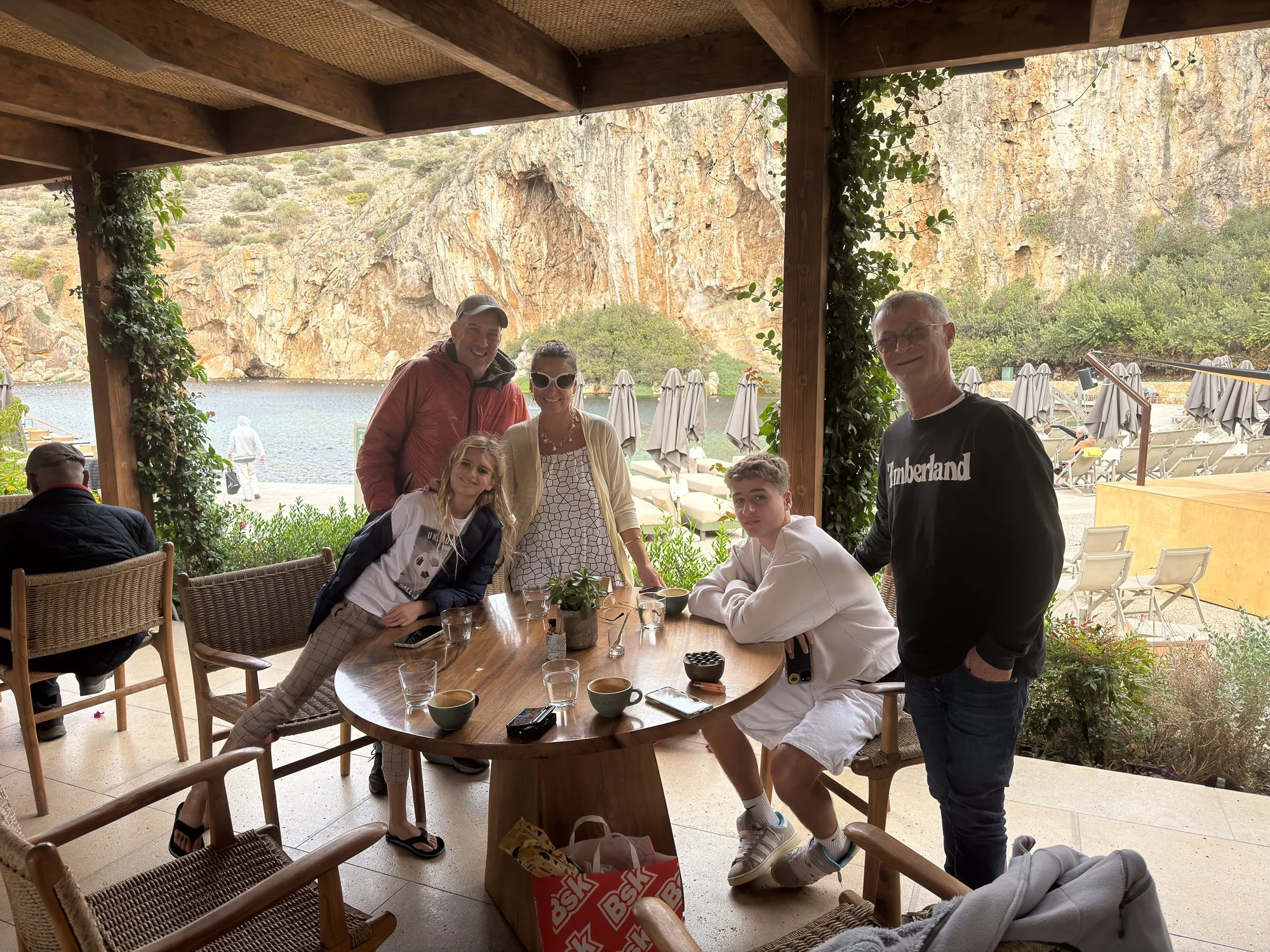 Group of six people at an outdoor patio with a view of a lake, mountains, and umbrellas on the beach in the background.