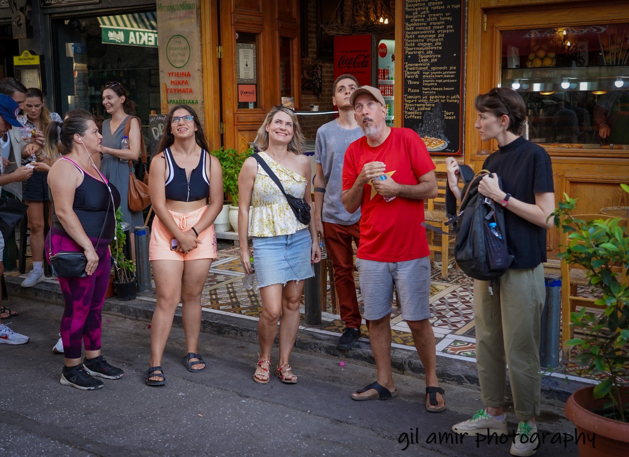 A group of people standing and waiting outside a restaurant on a city street. There are five people in the foreground, including men and women, some smiling and some looking around. In the background, others are walking and talking. The restaurant has a wooden exterior with a menu board and potted plants visible.
