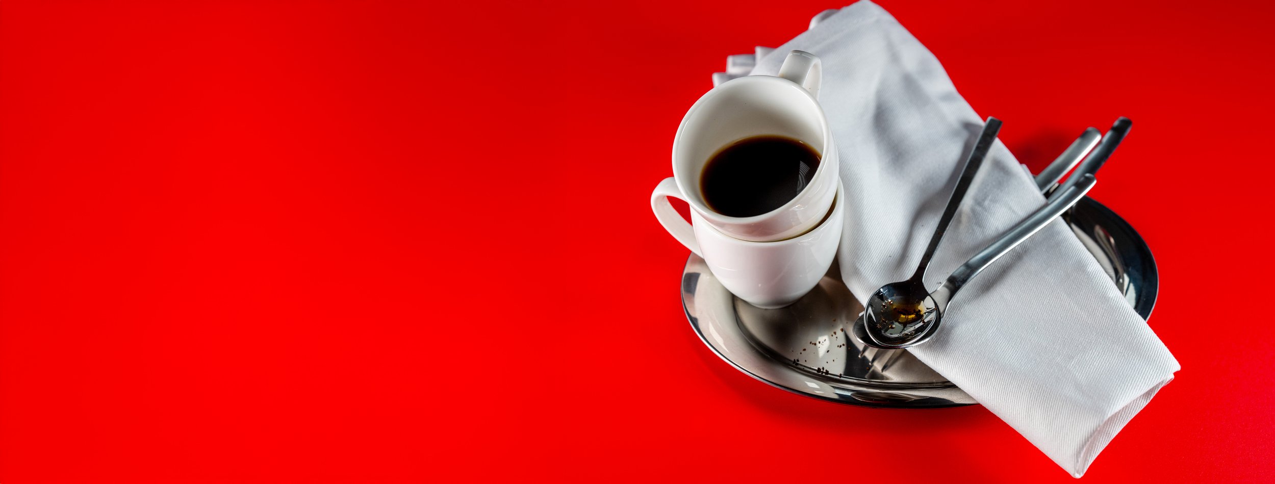 A metal tray holding a white coffee cup filled with black coffee, a white cloth napkin, and two silverware spoons on a bright red background.