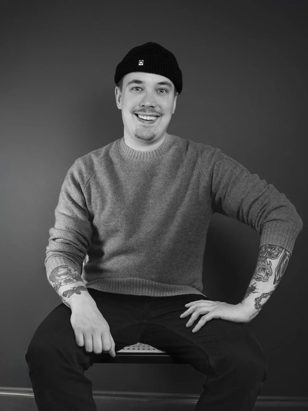 A young man with tattoos on his arms, wearing a knit cap and a sweater, sitting on a bench against a plain background, smiling at the camera.