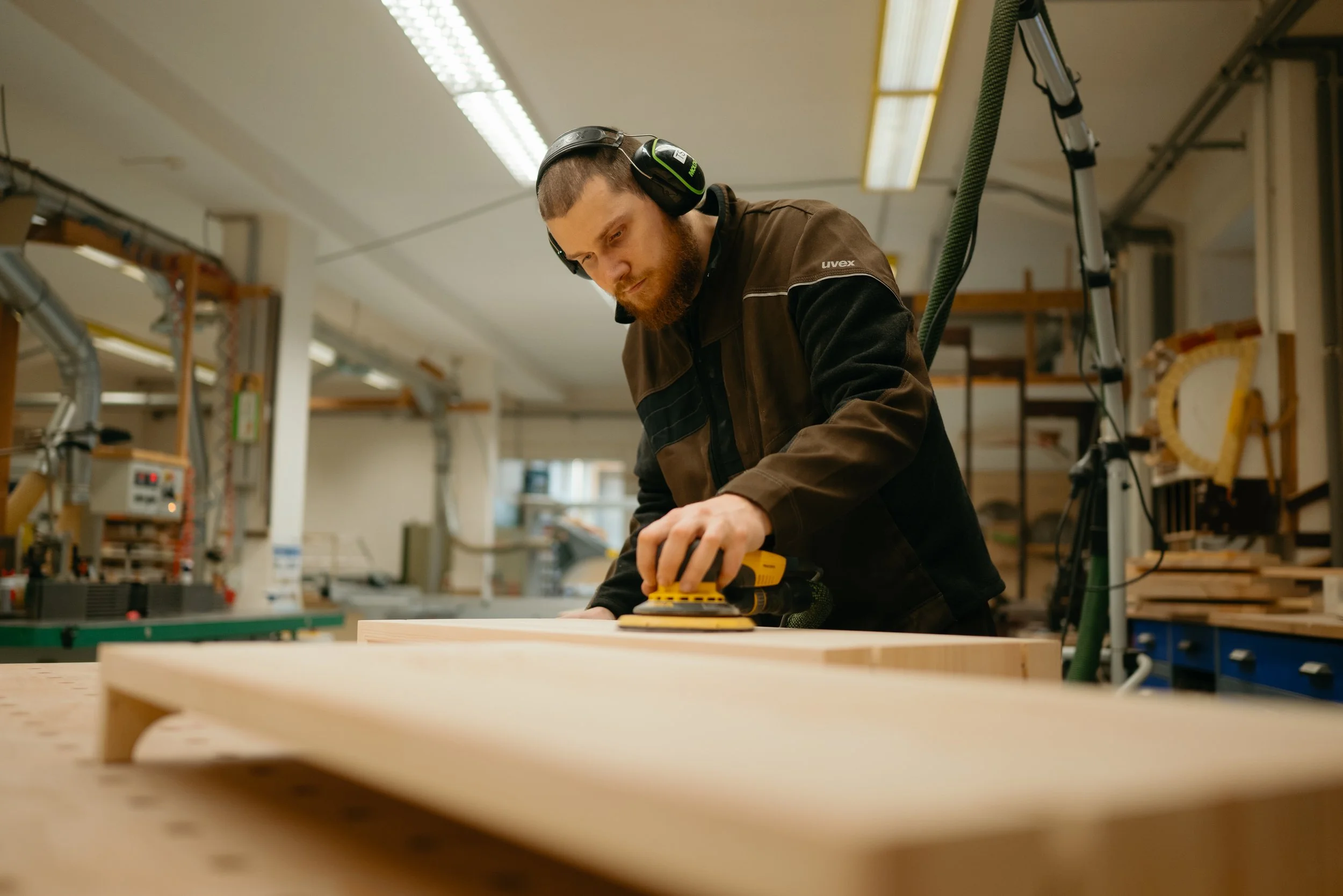 Mann in einer Werkstatt schleift ein Holzstück mit einem Elektroschleifer, trägt Gehörschutz, umgeben von Werkstatt-Equipment.