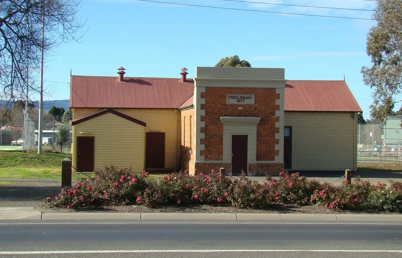 A small historic building labeled 'Free Library 1877' with a brick front and beige sides, surrounded by pink flowering bushes, with a clear blue sky and trees in the background located in  Wallan Victoria Australia