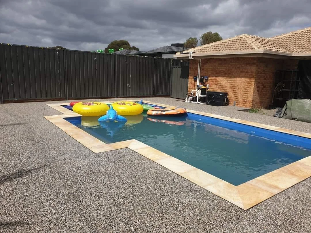 A backyard swimming pool filled with water, surrounded by a speckled concrete deck, with inflatable pool floats including yellow, blue, and orange, and a gray metal fence enclosures, under a cloudy sky.