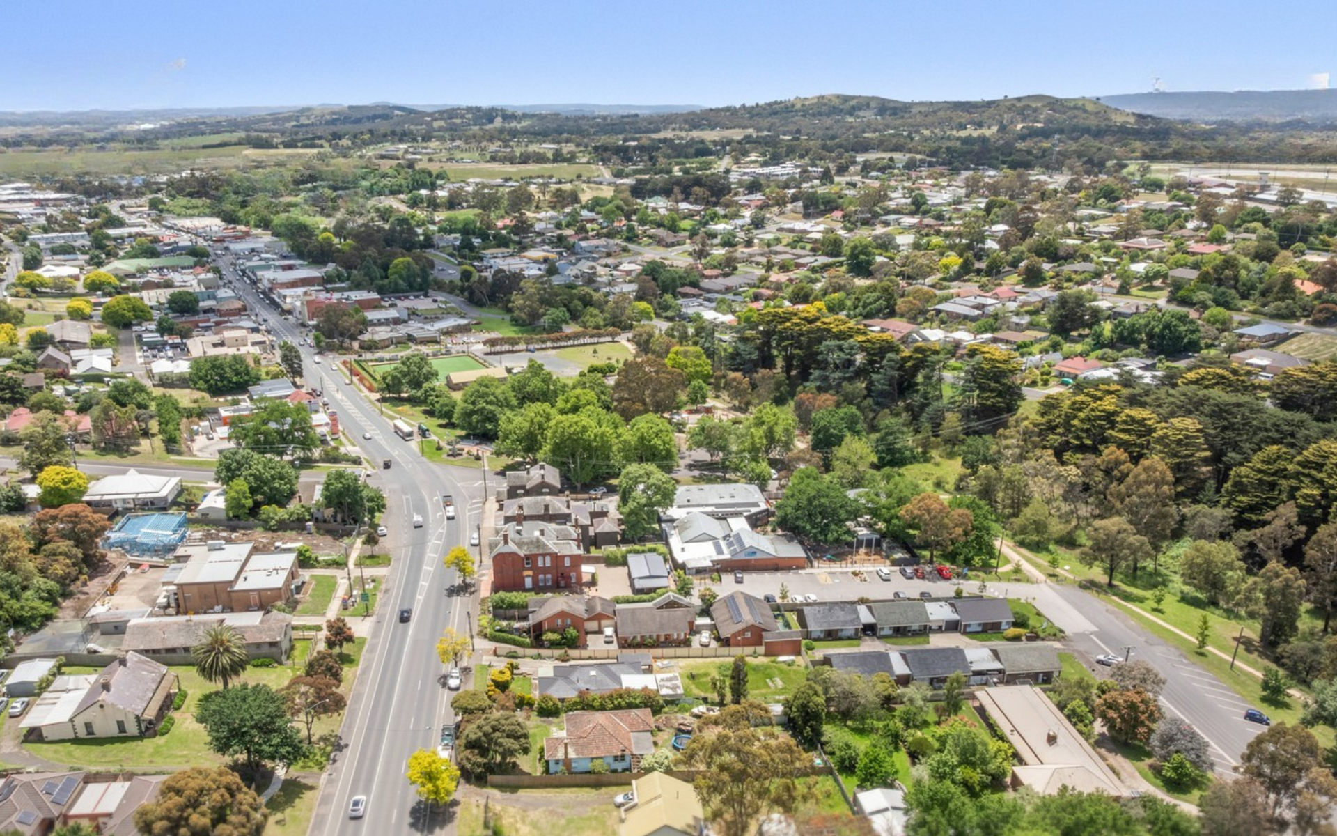 Aerial view of a suburban Kilmore Vic with houses, trees, and a main road running through the area, with hills in the background under a clear blue sky.