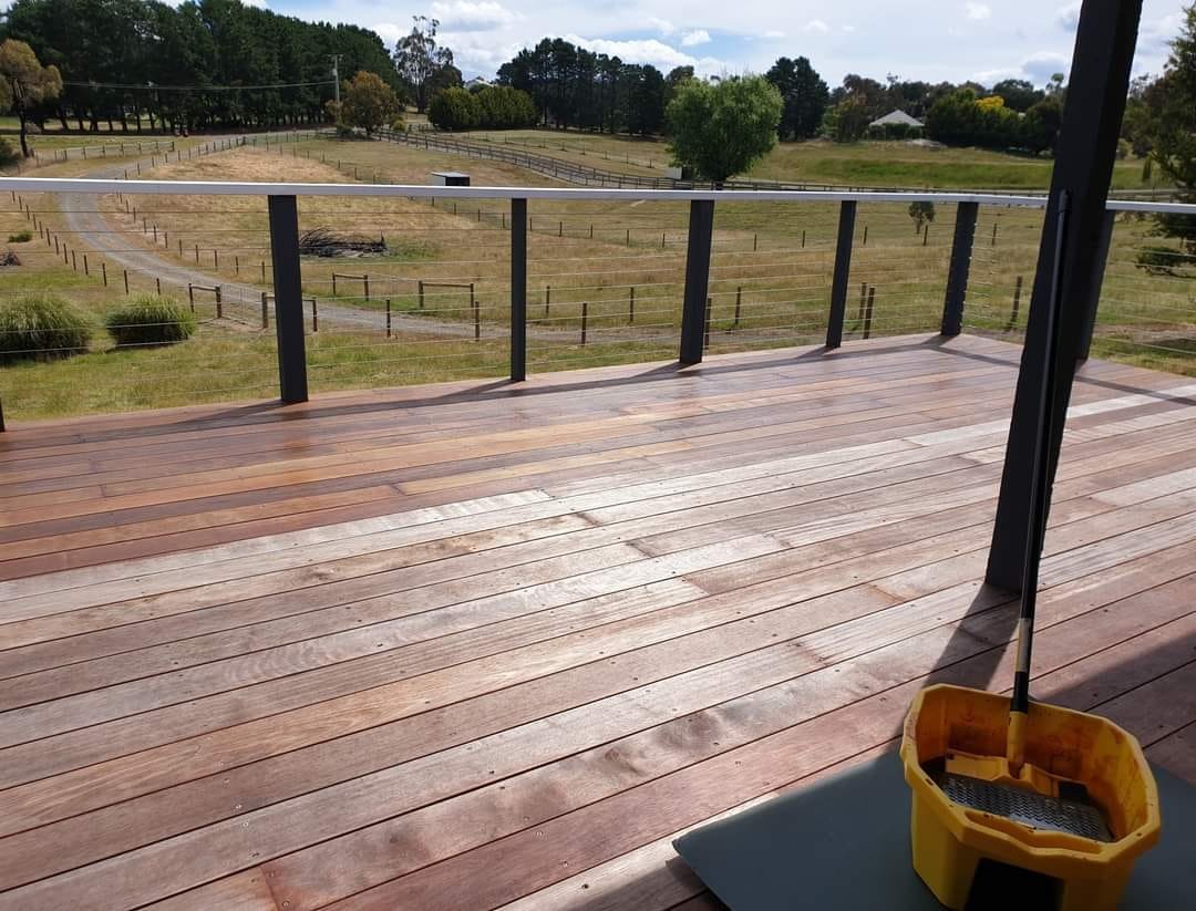 A wooden deck overlooking a grassy landscape with a winding dirt path, trees, and a cloudy sky in the background. A yellow mop bucket with a mop is on the deck in the foreground.