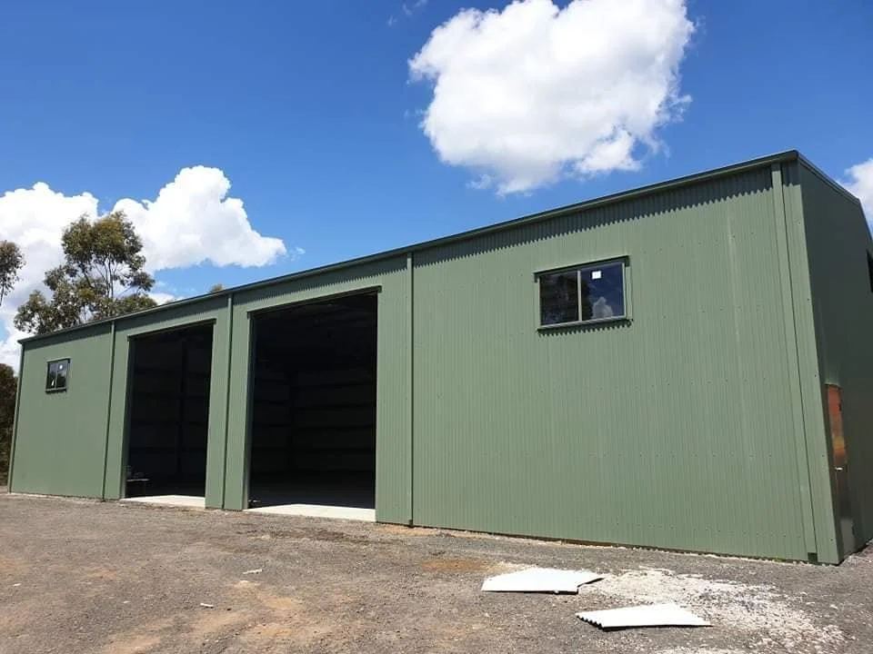 Green metal storage shed with two open bays and a small window on a gravel lot, under a partly cloudy sky.