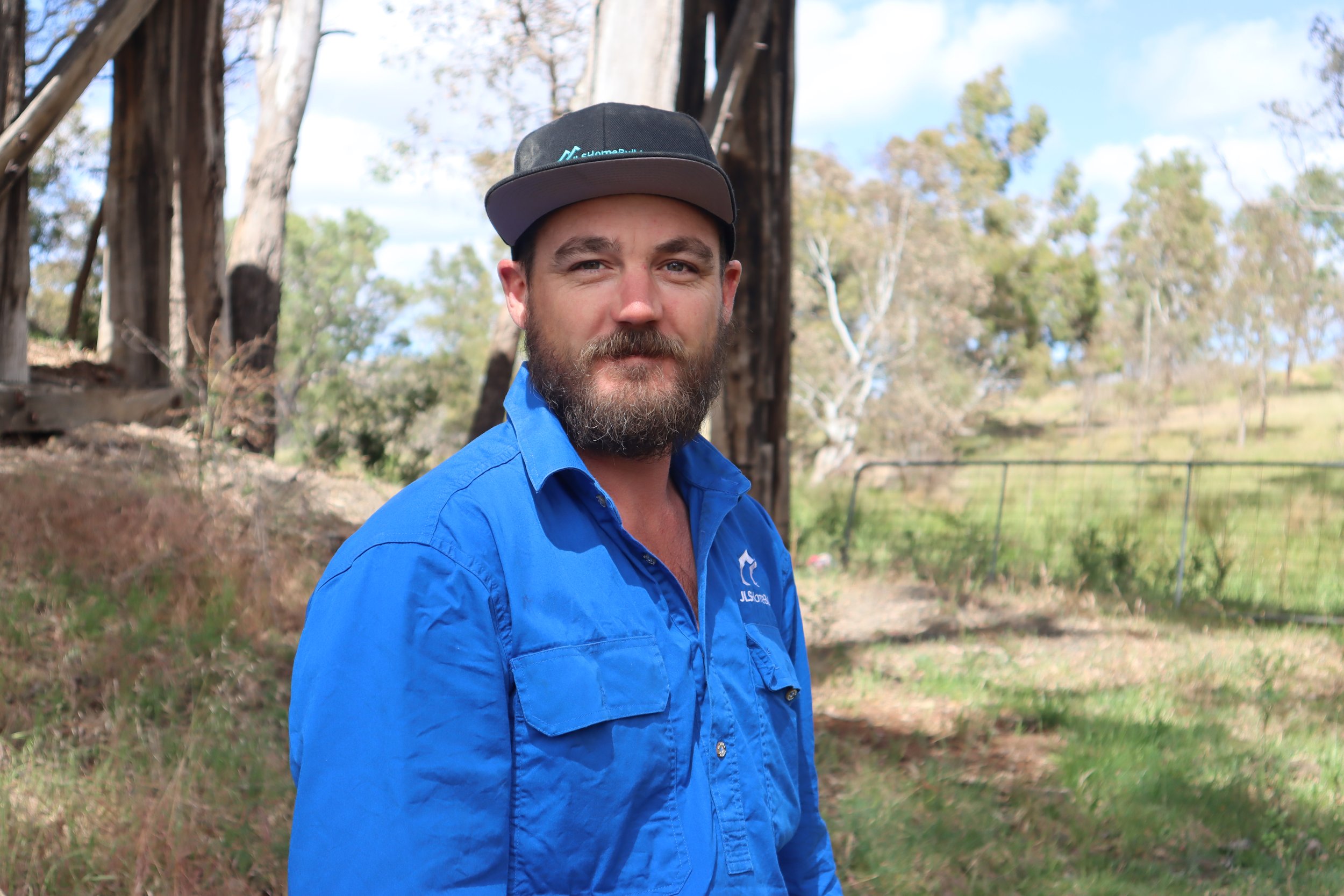 Jack standing in scenic rural country background, wearing a blue shirt and dark baseball hat with JLS Home Builders logo on his shirt and hat.
