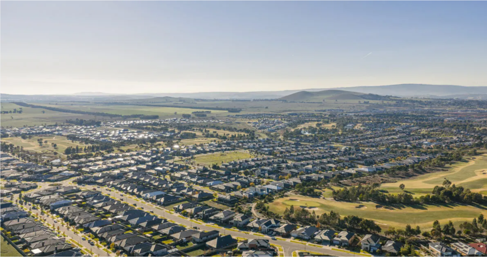 Aerial view of a residential neighborhood of Beveridge Victoria Australia with houses, streets, and a golf course in the foreground, with rolling hills in the background under a clear blue sky.