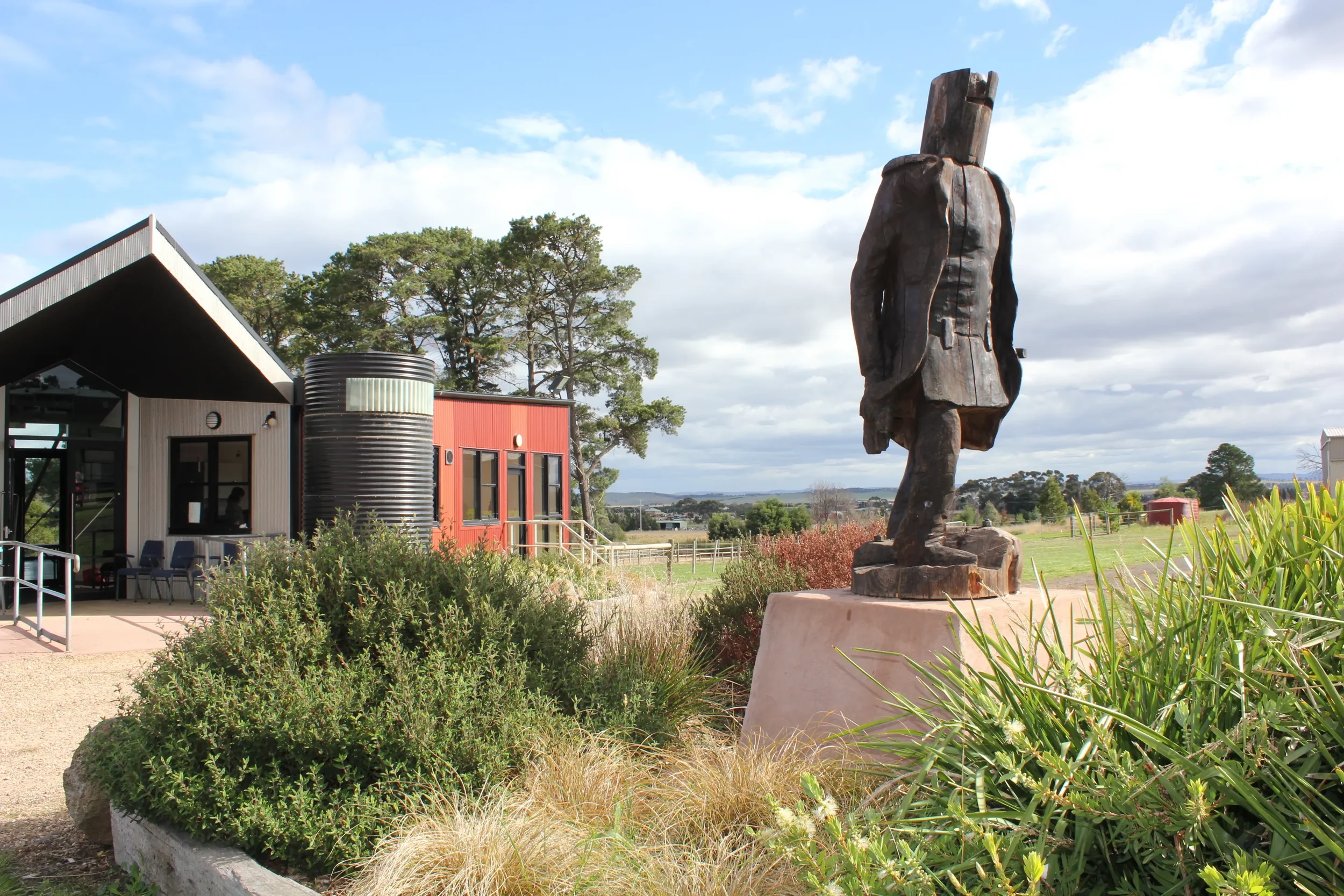 A landscaped outdoor area with a large wooden sculpture of NED KELLY, modern buildings with red and gray exteriors, and lush green trees under a partly cloudy sky located in Beveridge VIC Australia