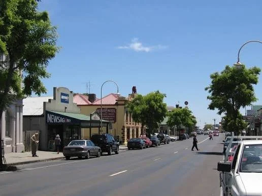 Street scene in a small town Kilmore Victoria Australia with parked cars, trees, and storefronts, including a news agency and other businesses, under a blue sky.