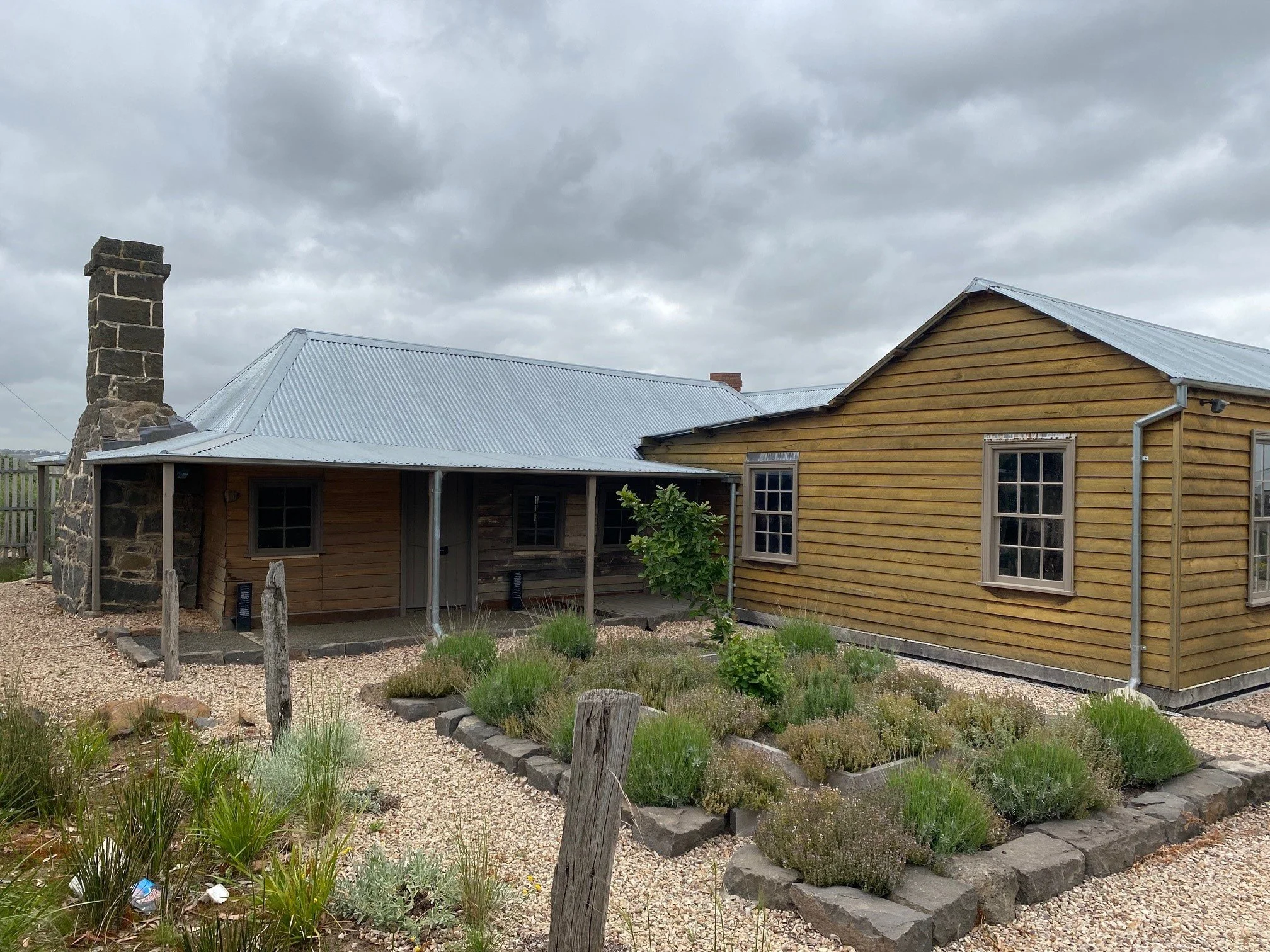 Photo of a house with a stone chimney of historic home in Beveridge VIC, surrounded by a garden with plants and a gravel pathway, under a cloudy sky.