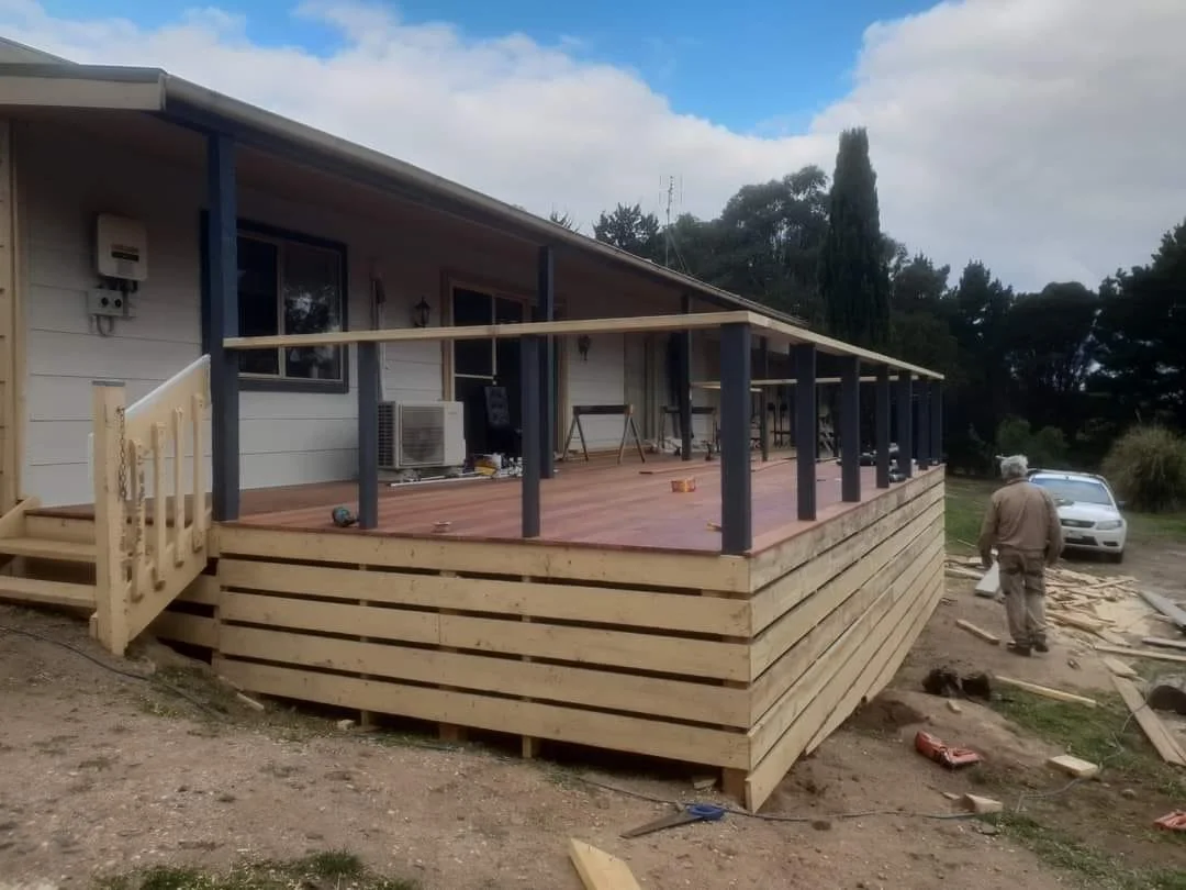 A house with a newly constructed wooden deck in progress, with tools and materials scattered around, and a worker standing nearby.