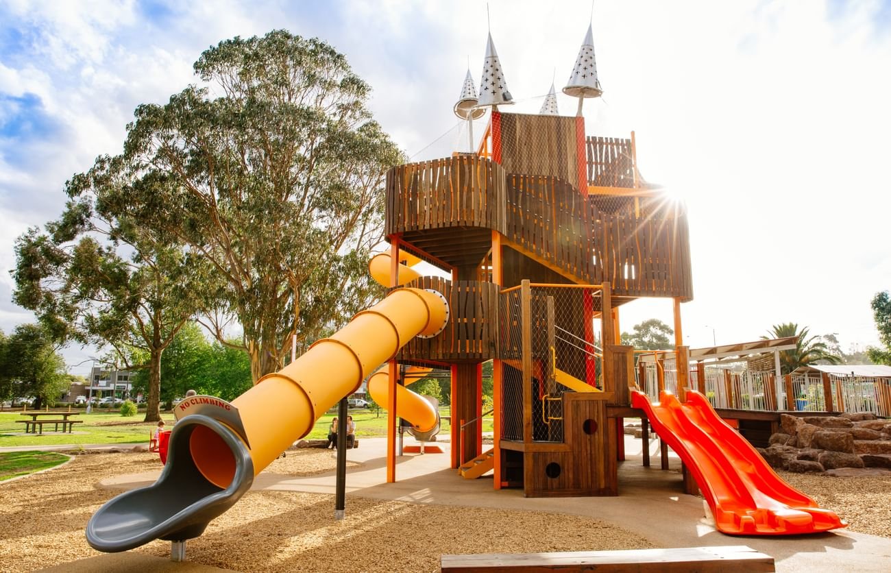 Children's playground structure located in Wallan VIC with a yellow tube slide, red side-by-side slides, and a tower with cone-shaped decorations, set in a park with trees and a blue sky.