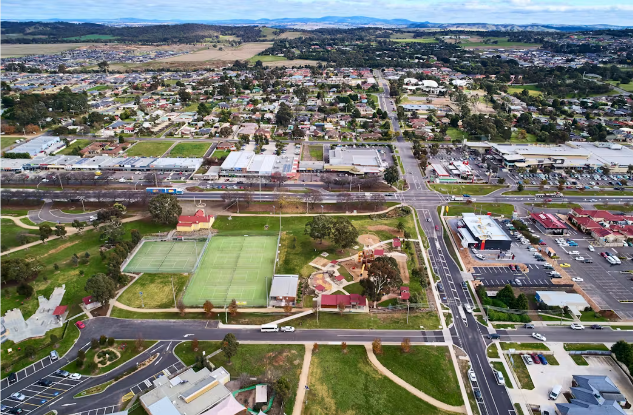 An aerial view of a small town Wallan VIC showing a mix of residential houses, commercial buildings, parking lots, and a park with tennis courts and playgrounds. Roads and streets are visible, with some cars traveling along them.