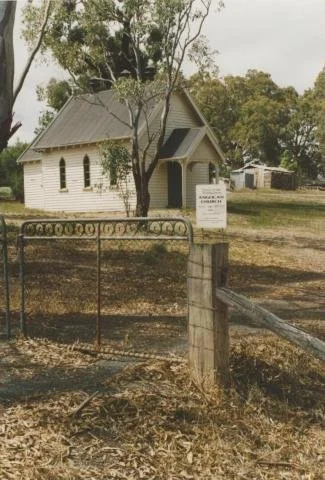 A small white church in Pyalong Victoria Australia with a steeple and arched windows behind a rusted iron gate and a weathered wooden fence, with trees and a cloudy sky in the background.