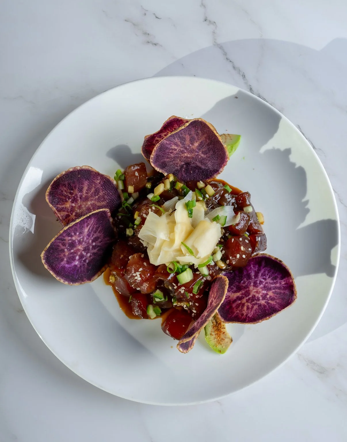 A white plate with sashimi-style raw fish topped with green onions, ginger, sesame seeds, and surrounded by purple potato chips, all on a white marble surface.