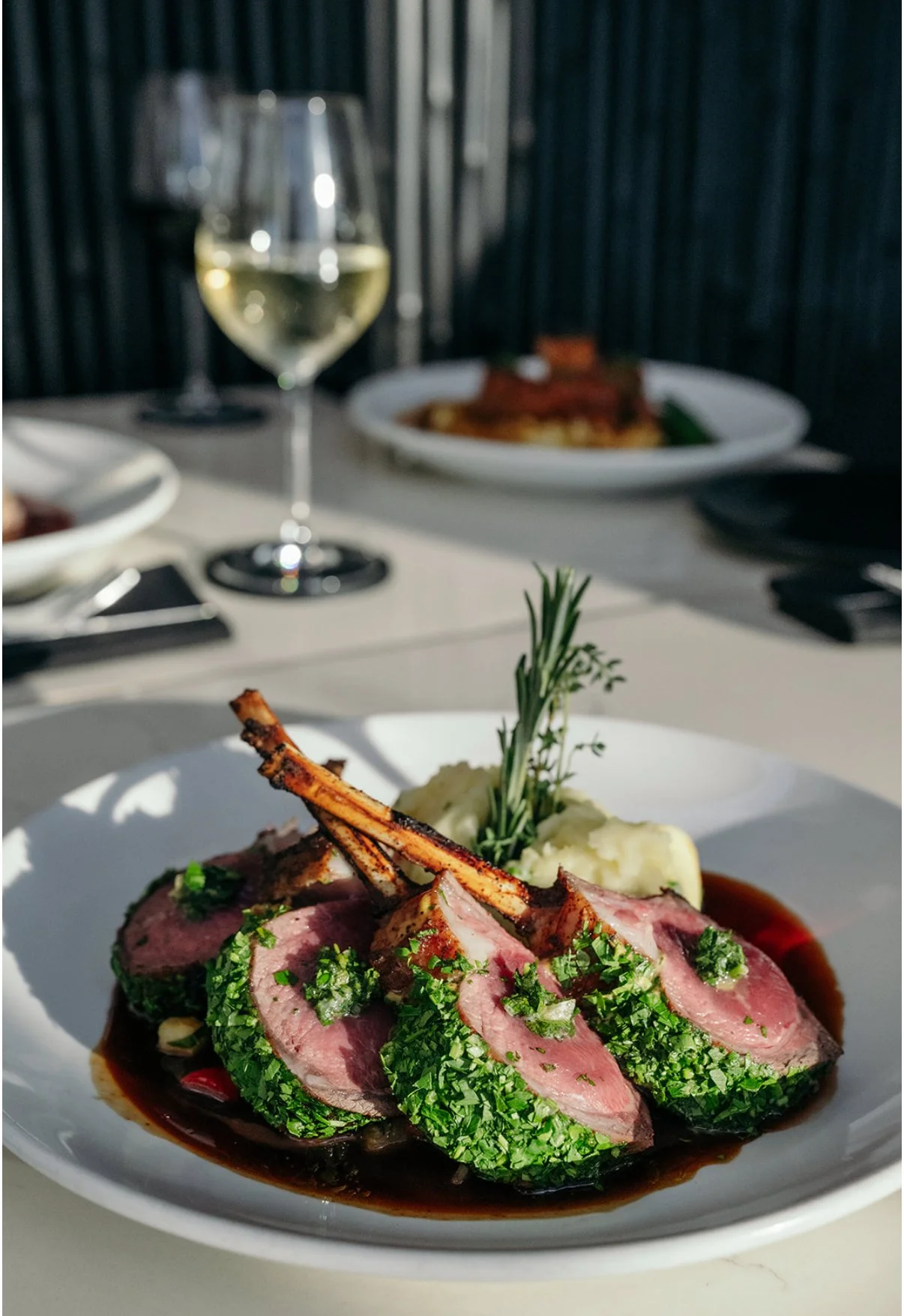 A plate of sliced lamb with herbs, mashed potatoes, and a sprig of rosemary, accompanied by a glass of white wine on a table in a well-lit restaurant.