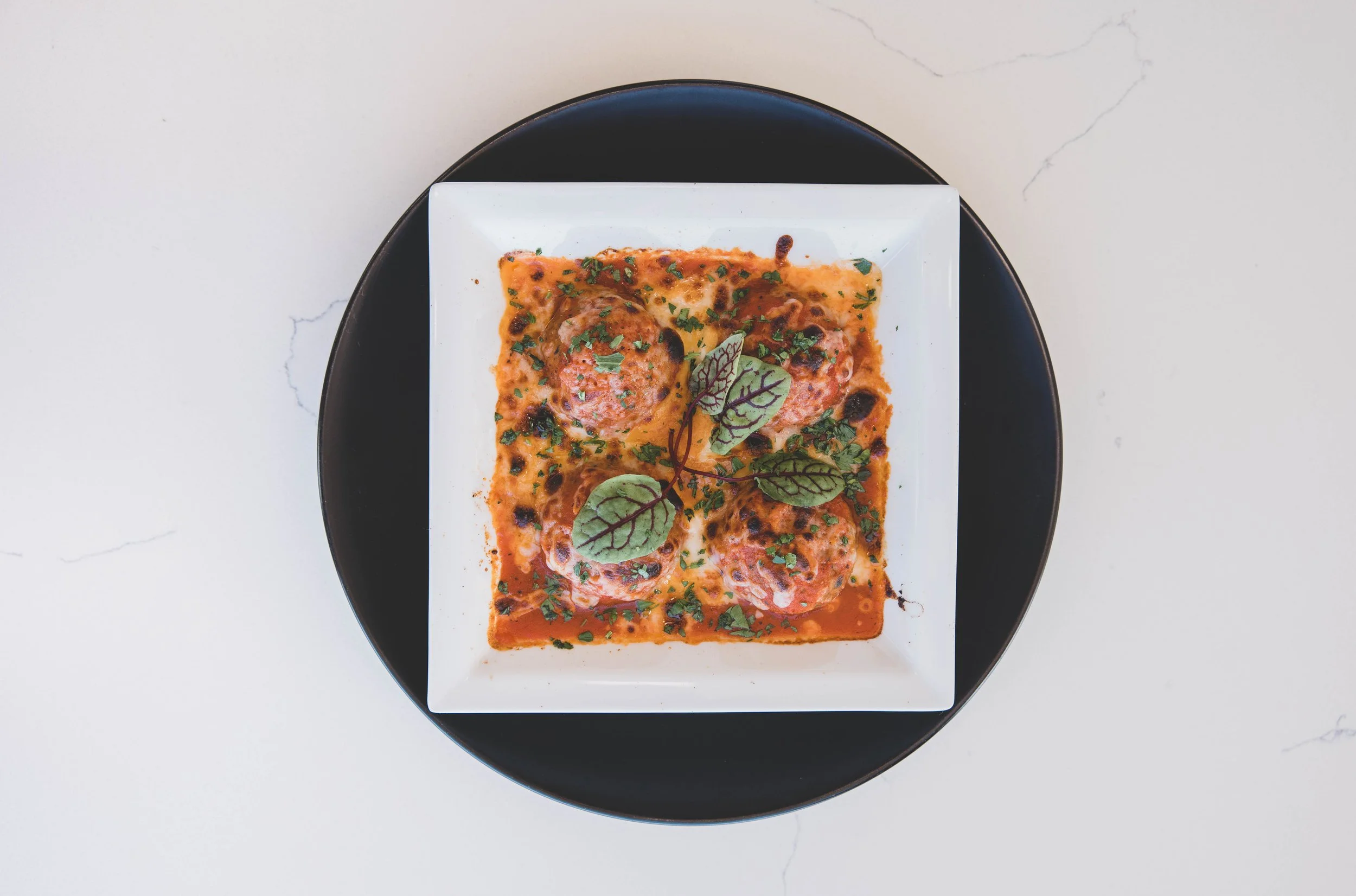 A square white plate with four meatballs in a red sauce garnished with green herbs and a sprig of purple-veined greens, on a black round tray.