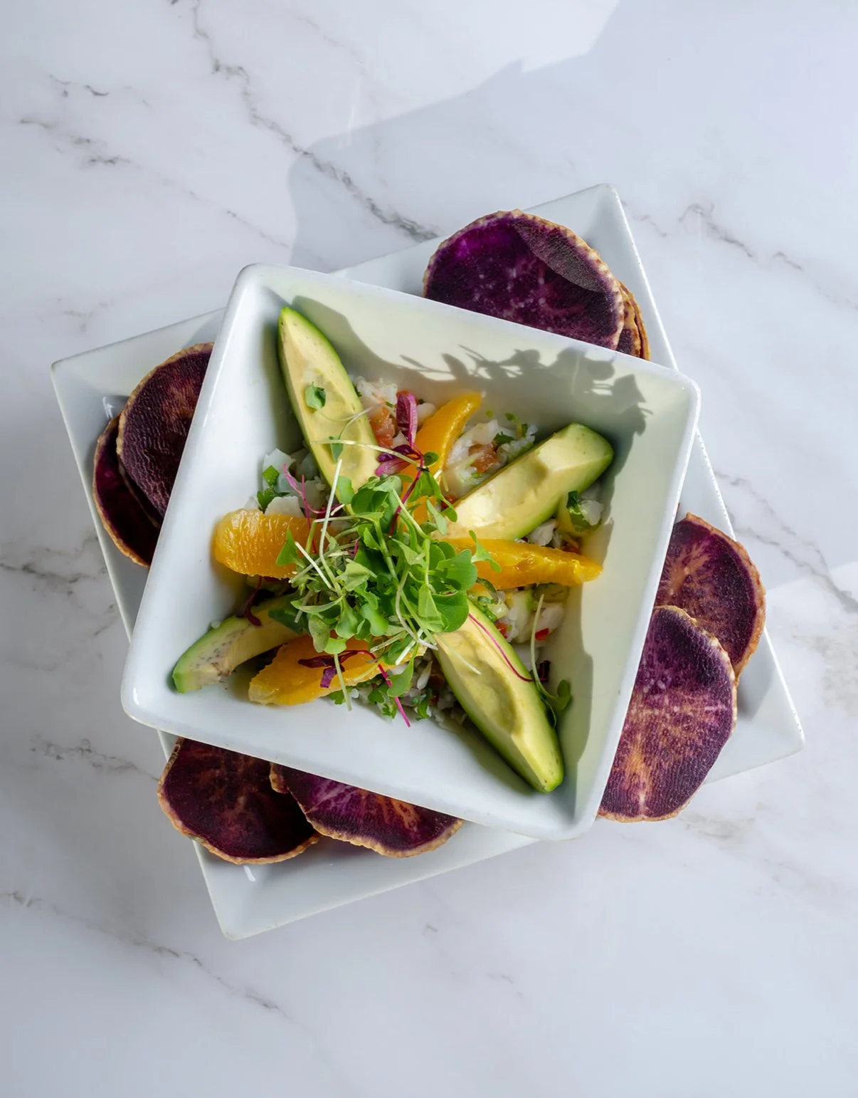A colorful salad with avocado, orange segments, and microgreens in a white bowl, garnished with purple sweet potato chips on a square white plate on a marble surface.