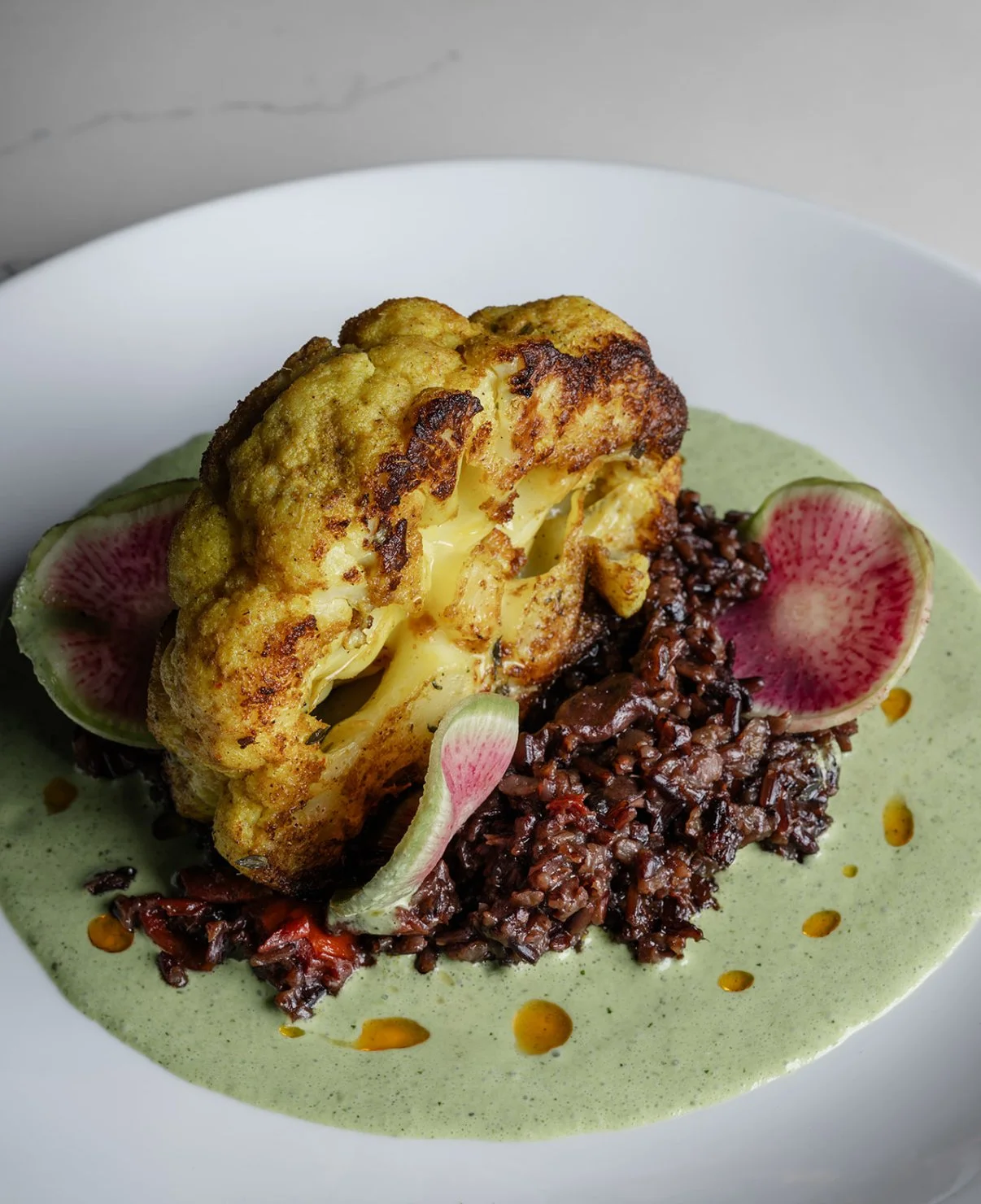 A plate of roasted cauliflower atop a bed of black rice, garnished with watermelon radish slices and drizzled with green sauce and oil droplets.
