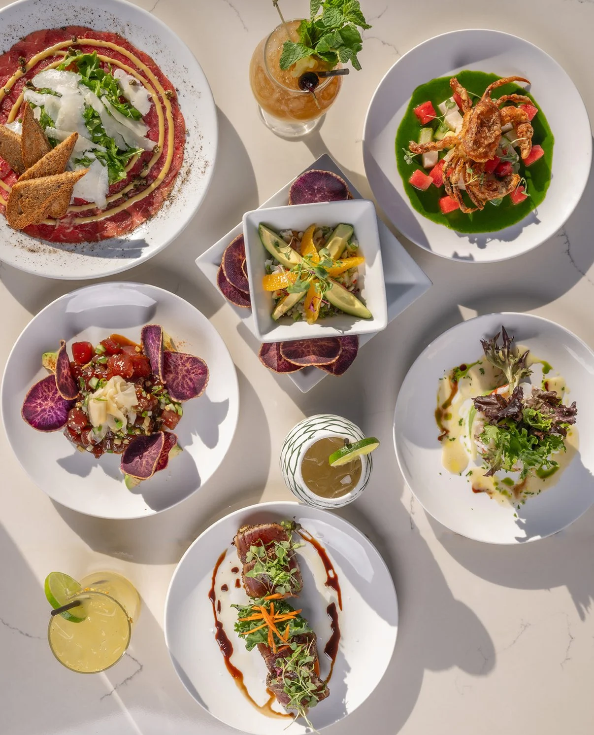 A variety of gourmet dishes on white plates, including salads, a sliced meat dish with sauce, a seafood dish with green sauce, topped with edible flowers, sliced vegetables, and microgreens, along with two cocktails garnished with lime and mint, all 