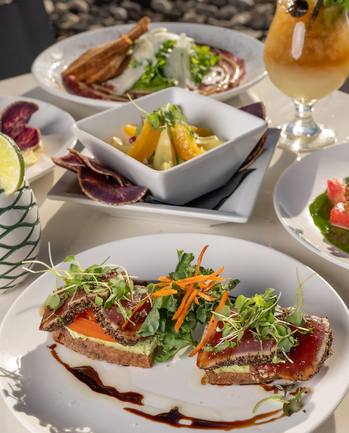 Appetizer plate with slices of toast topped with cured meat, avocado, and microgreens, garnished with leaf and shredded carrot, served with balsamic glaze.