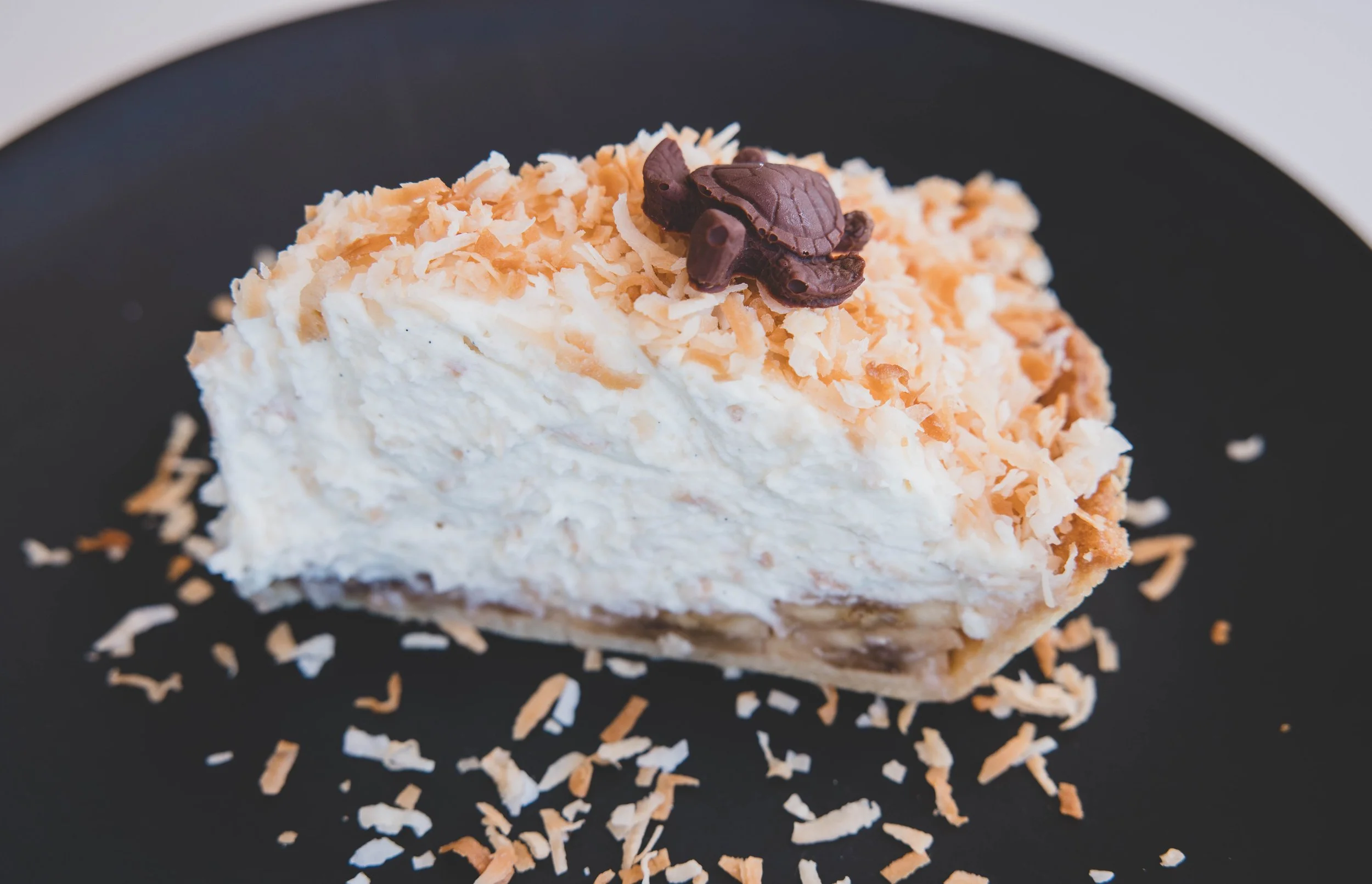 Slice of coconut cream pie topped with shredded coconut and a chocolate turtle on a black plate.
