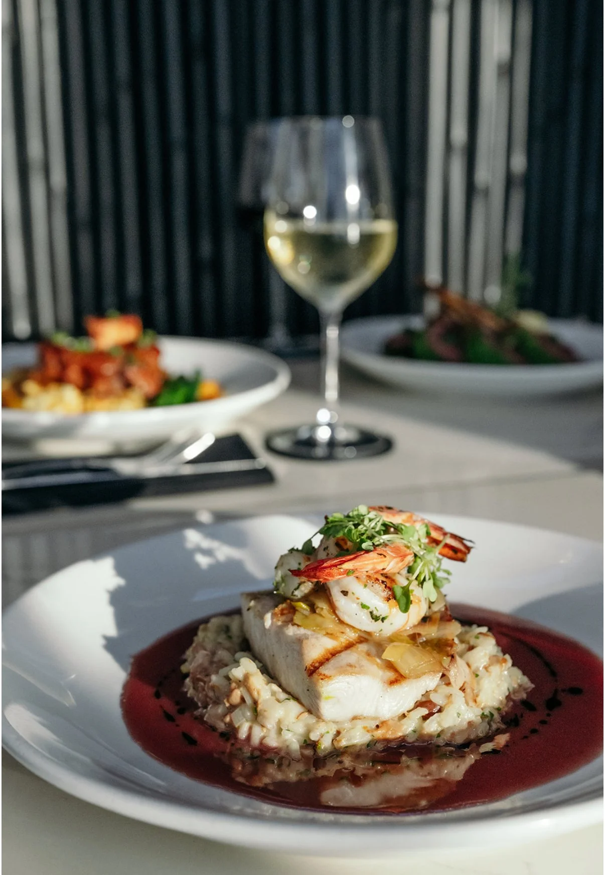 A plated seafood dish featuring a white fish fillet topped with shrimp and greens, served over a creamy risotto with a red sauce on a white plate. In the background, there is a glass of white wine and additional plates with food on a white tablecloth