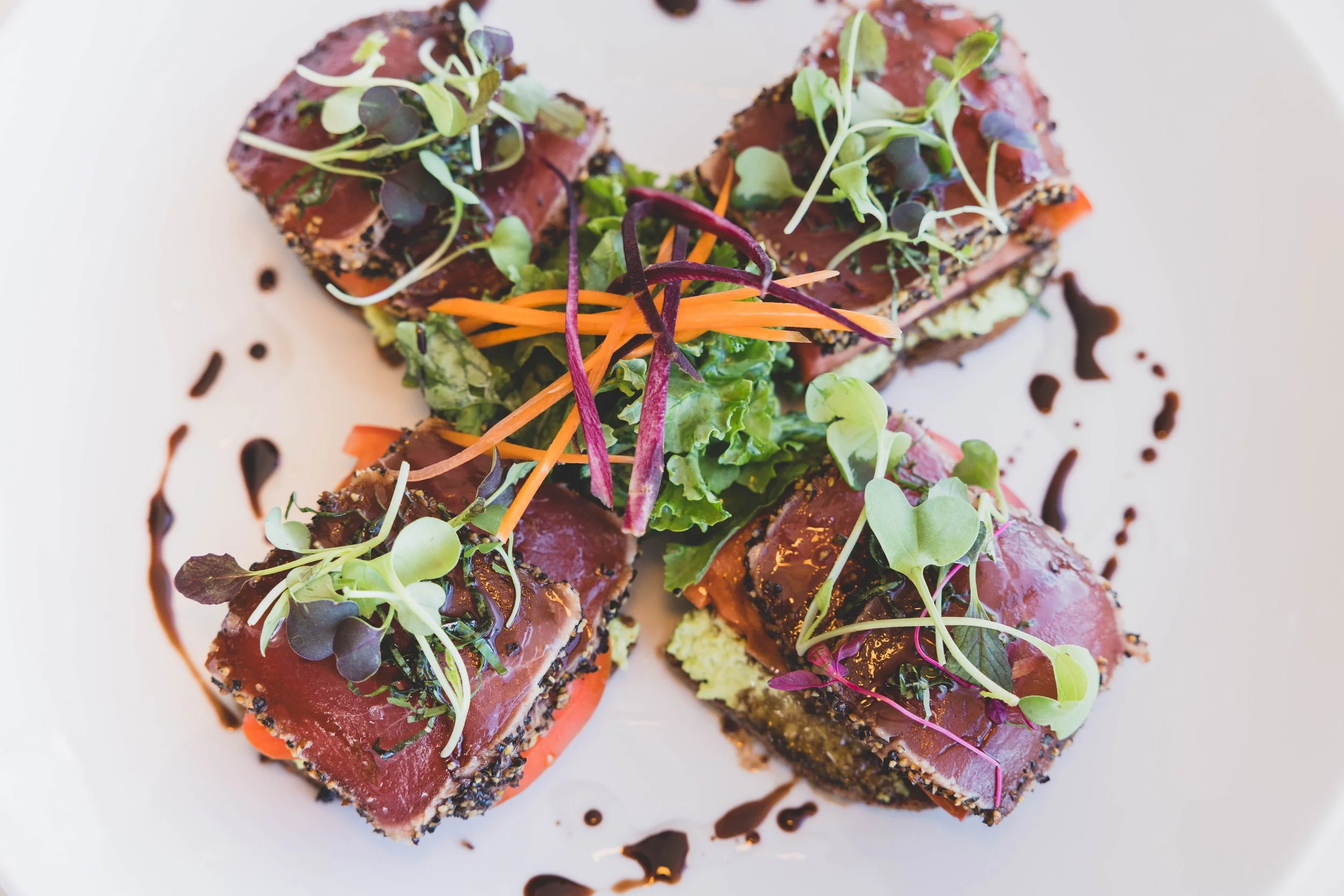 Seared tuna slices topped with microgreens, served with shredded carrots and purple cabbage, garnished with a drizzle of balsamic glaze on a white plate.