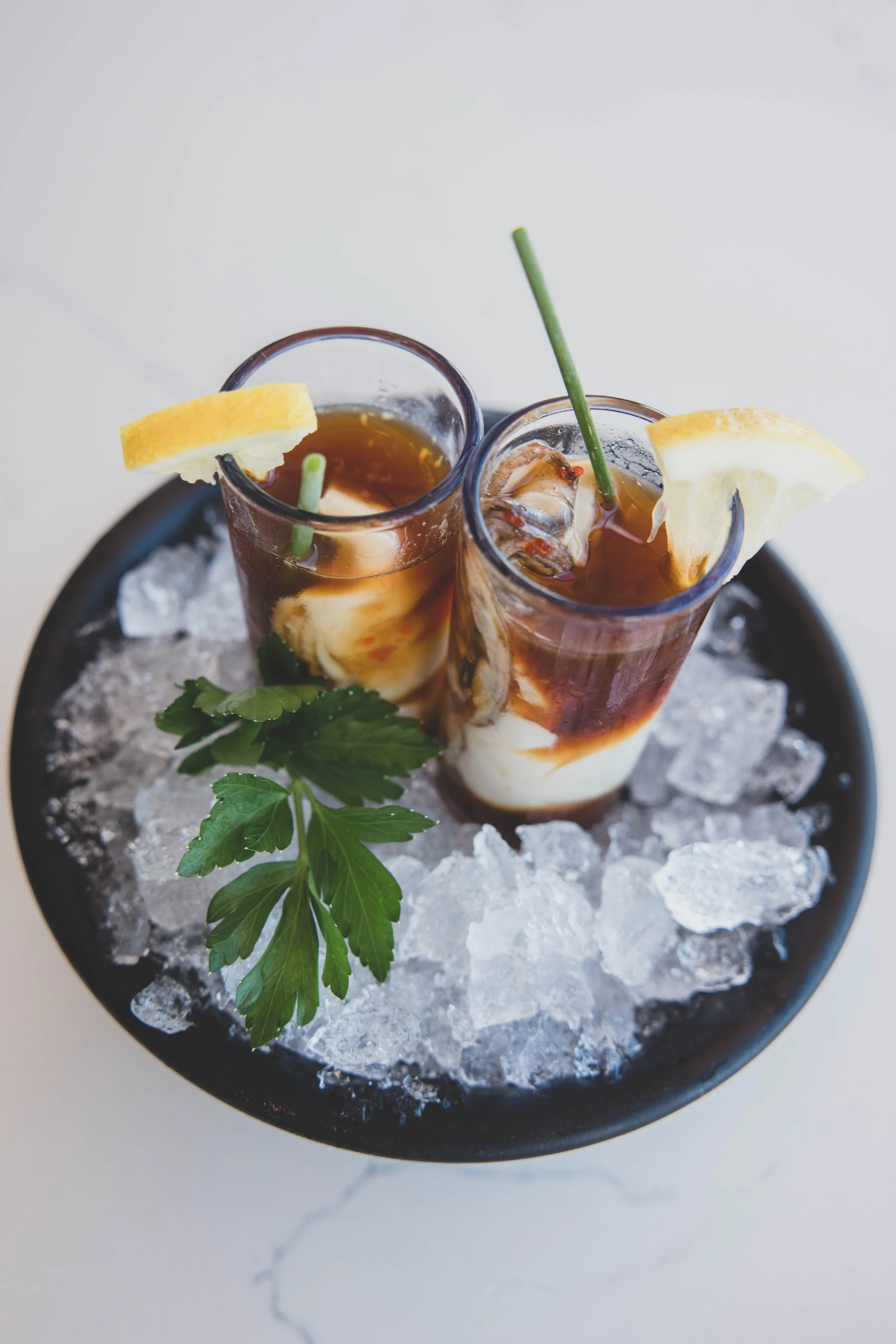 Two glasses of iced tea with lemon wedges and straws, garnished with a sprig of parsley, placed on a bed of ice in a black bowl.