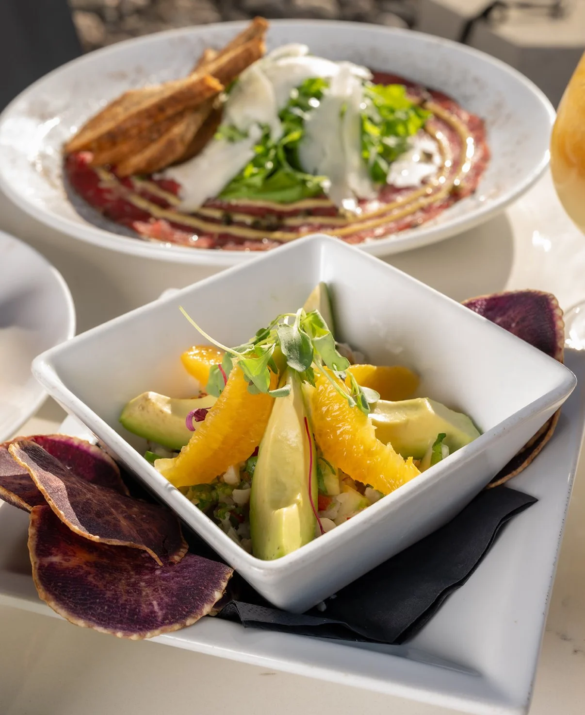 A bowl of fresh salad with orange slices, avocado, and microgreens, accompanied by purple plantain chips, in an elegant restaurant setting.
