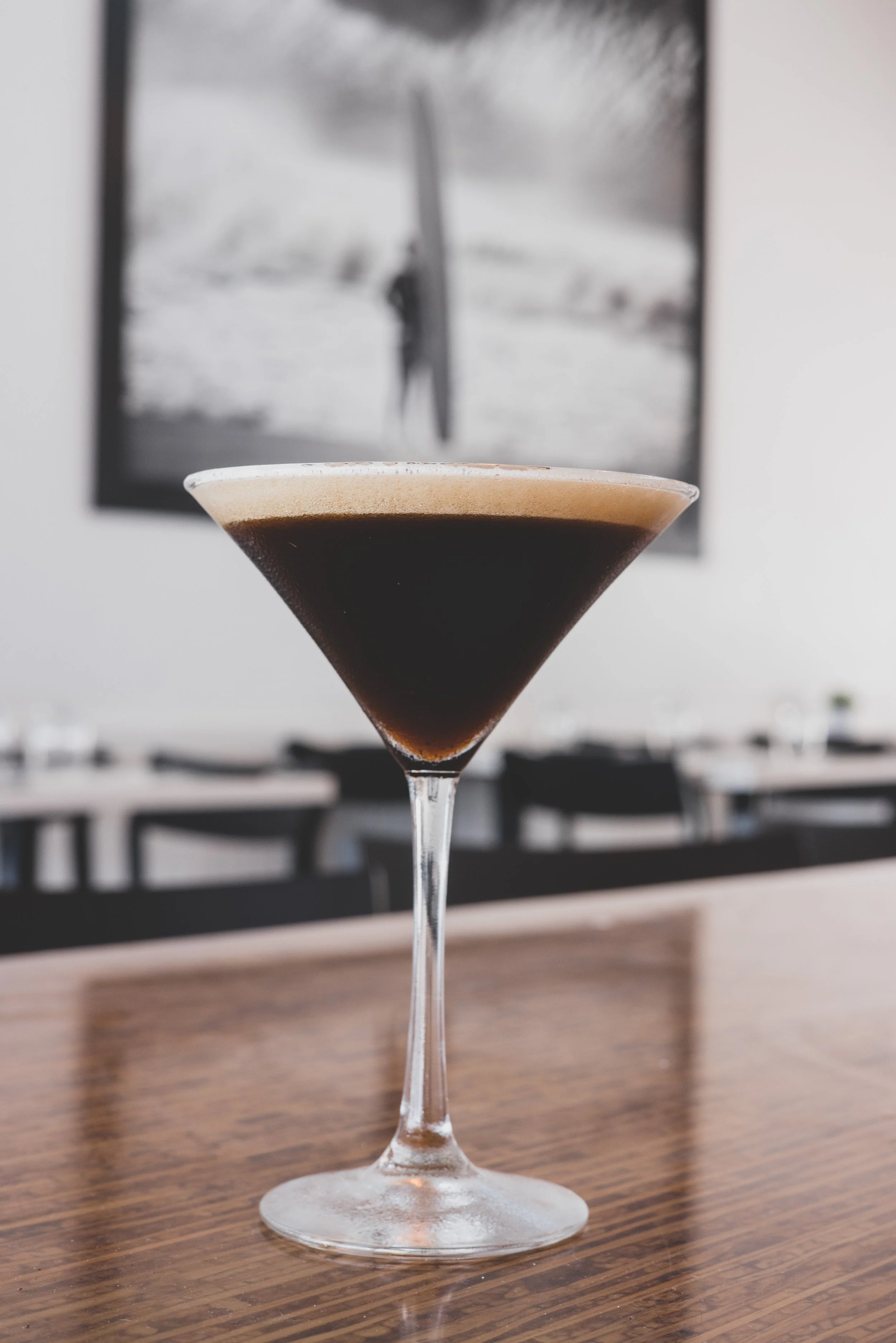 A dark cocktail in a martini glass on a wooden table with black chairs and a black-and-white photo in the background.